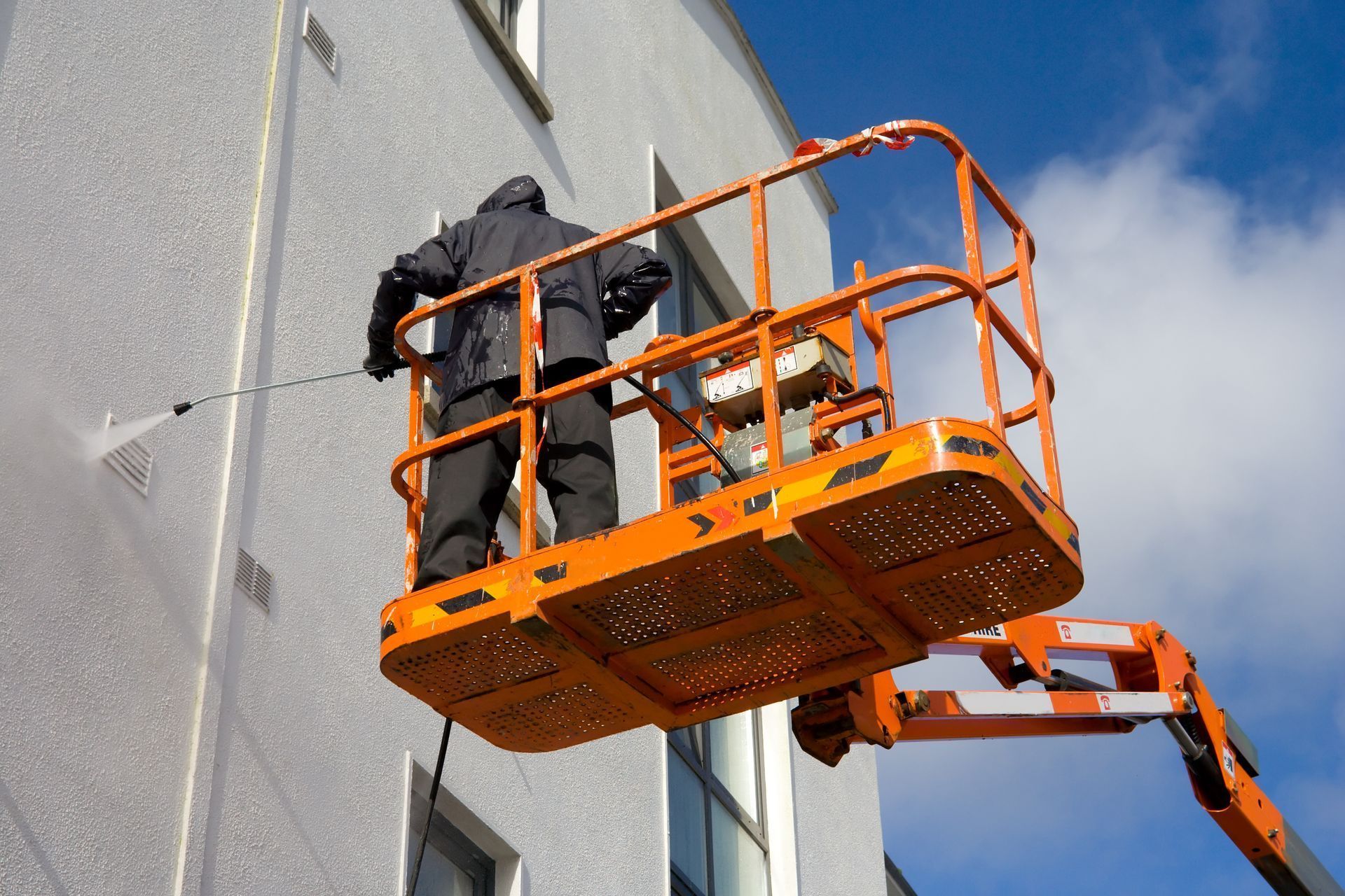 A worker in dark clothing uses a pressure washer to clean a white building wall from an orange aerial work platform.