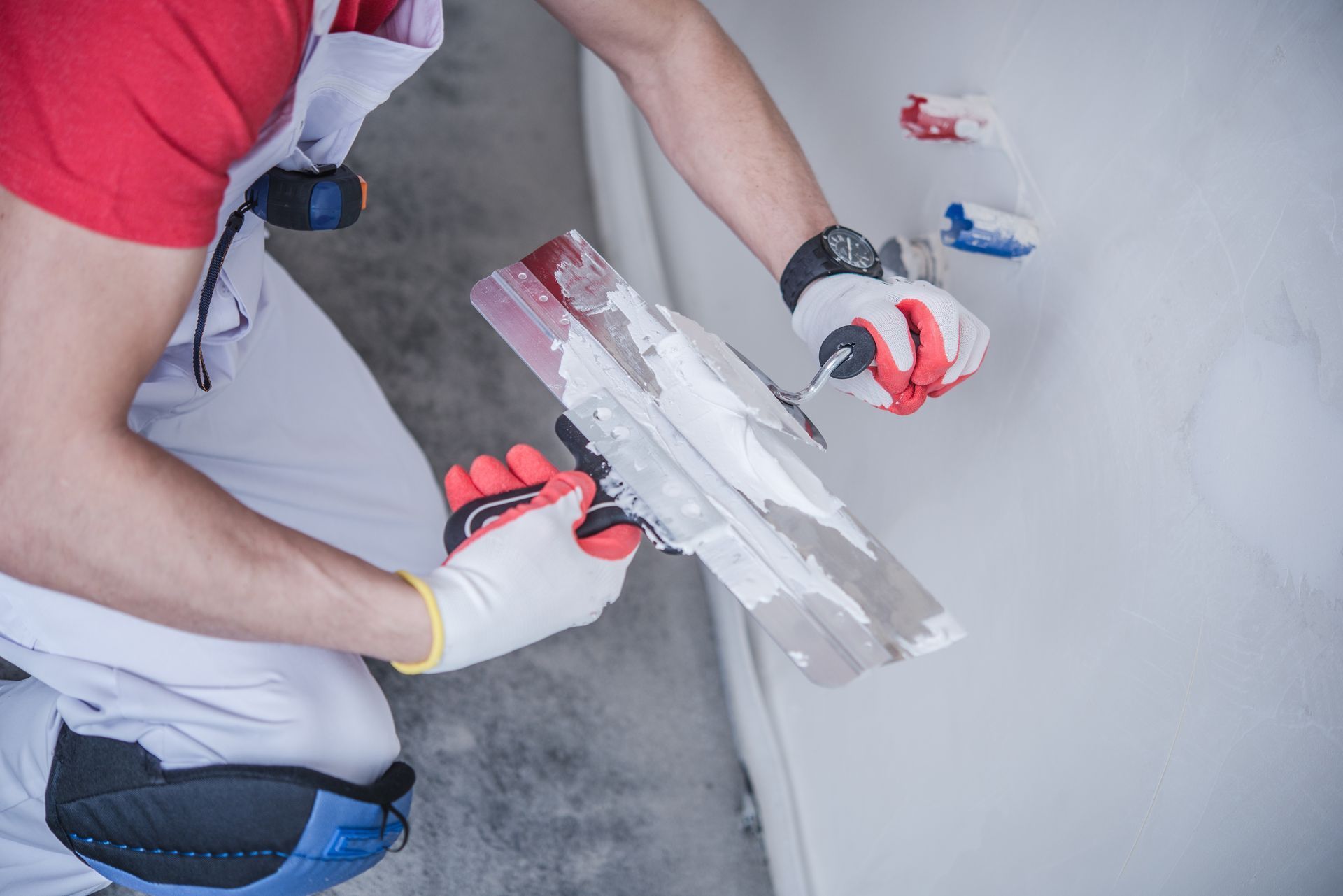 A construction worker in a red shirt and white overalls uses a plastering trowel to smooth white filler onto a wall.