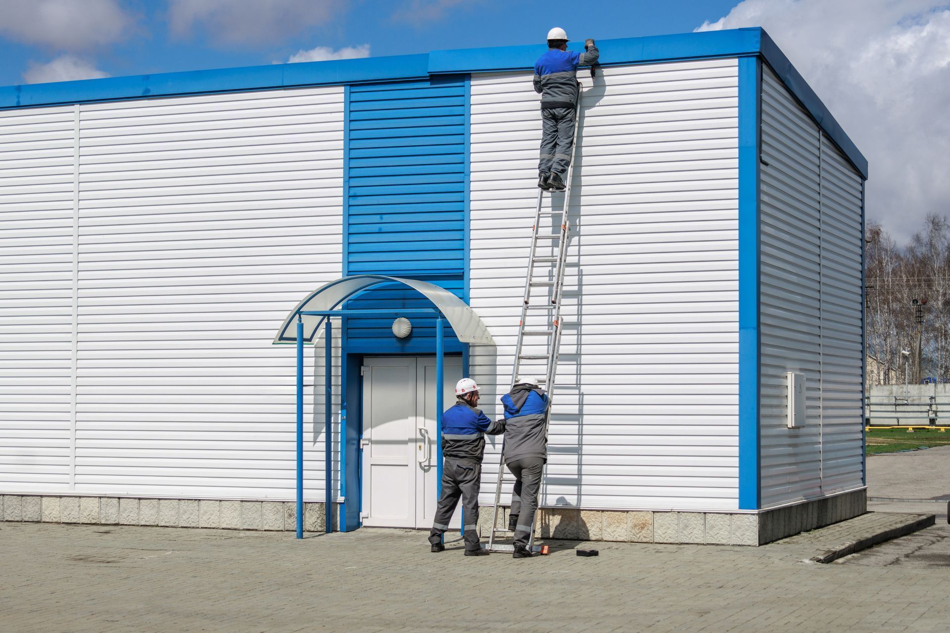 Three workers in uniforms and hard hats perform maintenance on a white and blue industrial building using a tall ladder.