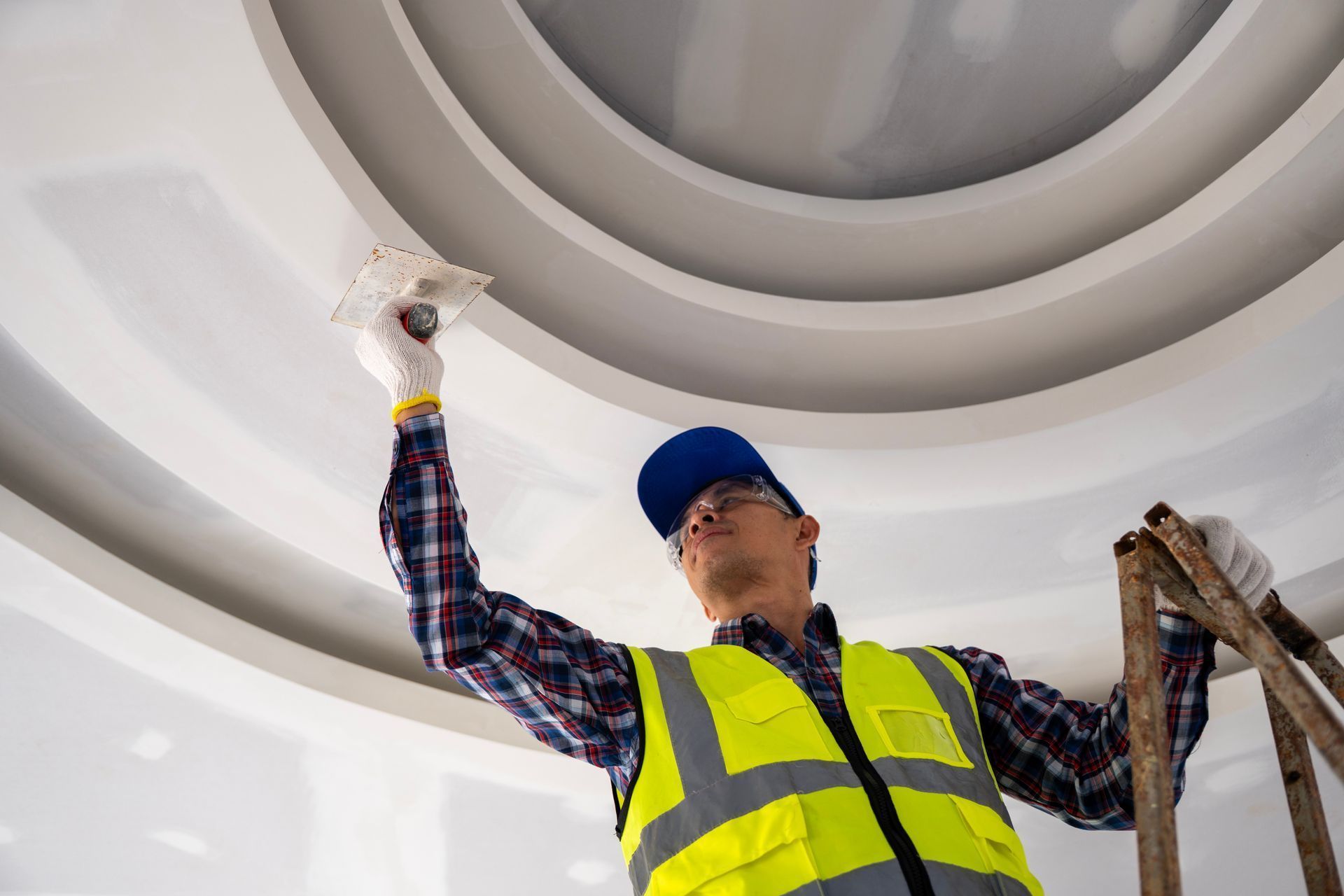 A worker in a yellow reflective vest and blue cap uses a tool to smooth drywall on a circular ceiling feature.