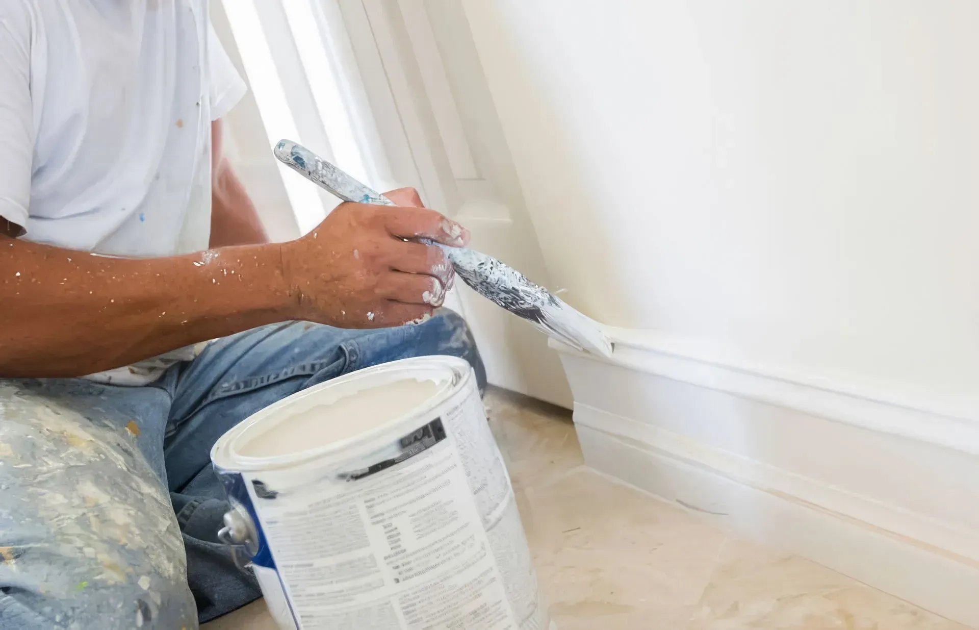 A person kneeling on the floor while painting white baseboard trim with a brush and a can of white paint.