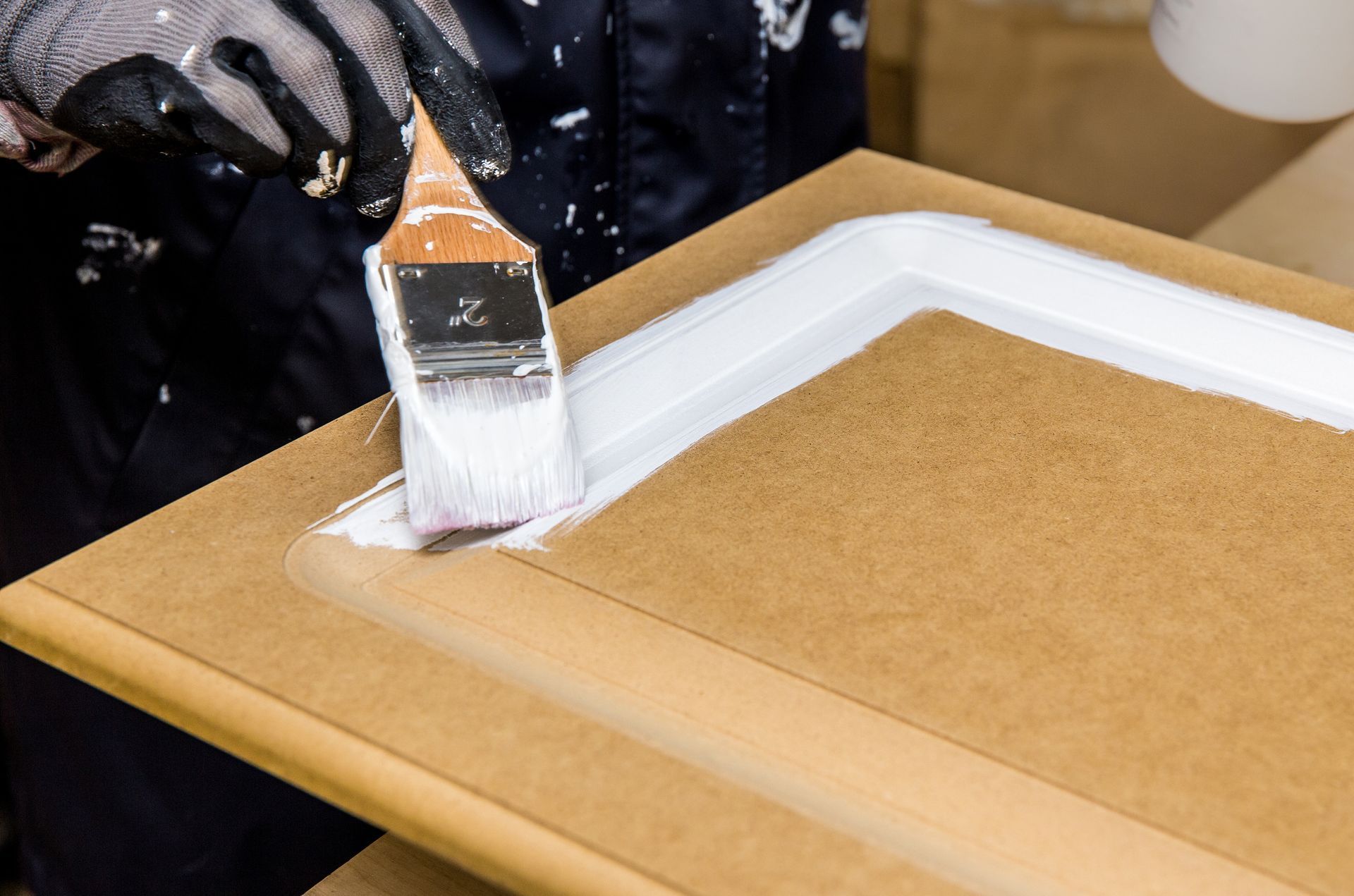 A person wearing work gloves uses a paintbrush to apply white paint to a wooden cabinet door.