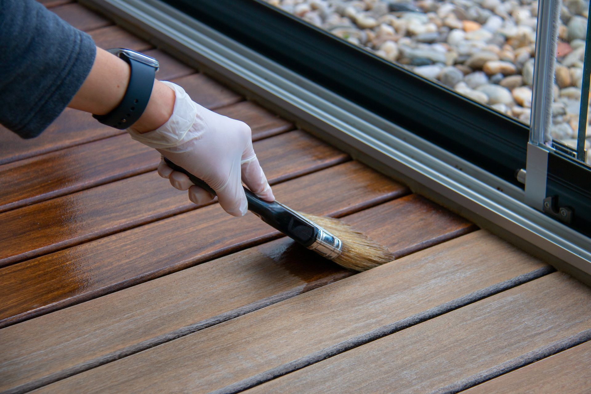 A hand in a white glove uses a brush to apply dark wood stain to brown deck planks beside a sliding glass door.