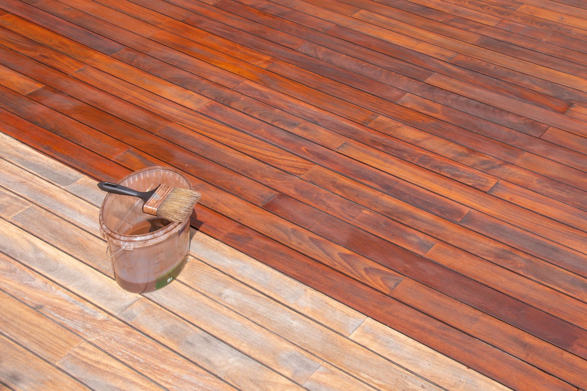 A brush in a paint bucket rests on a wooden deck, showing the contrast between unfinished and stained, dark wood planks.