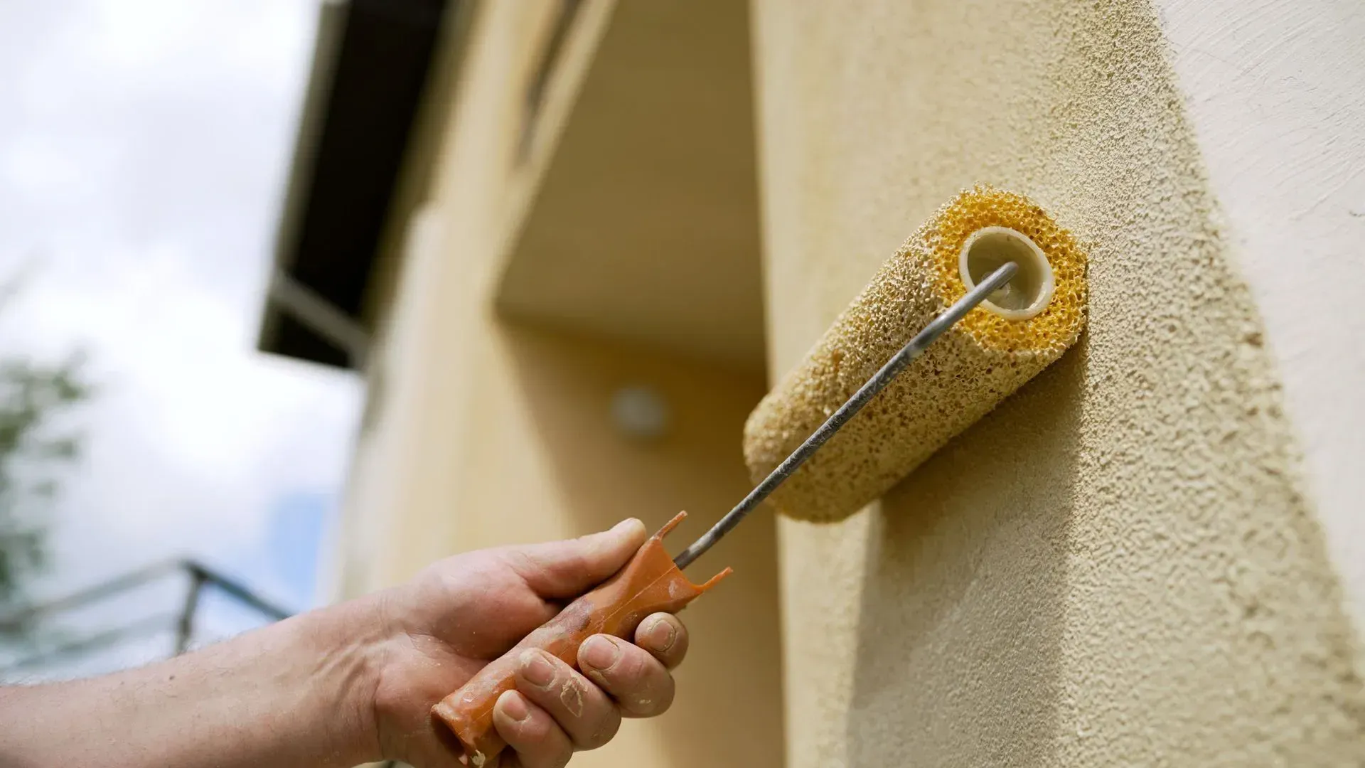 A close-up of a hand holding a textured paint roller, applying beige paint to a rough exterior wall.