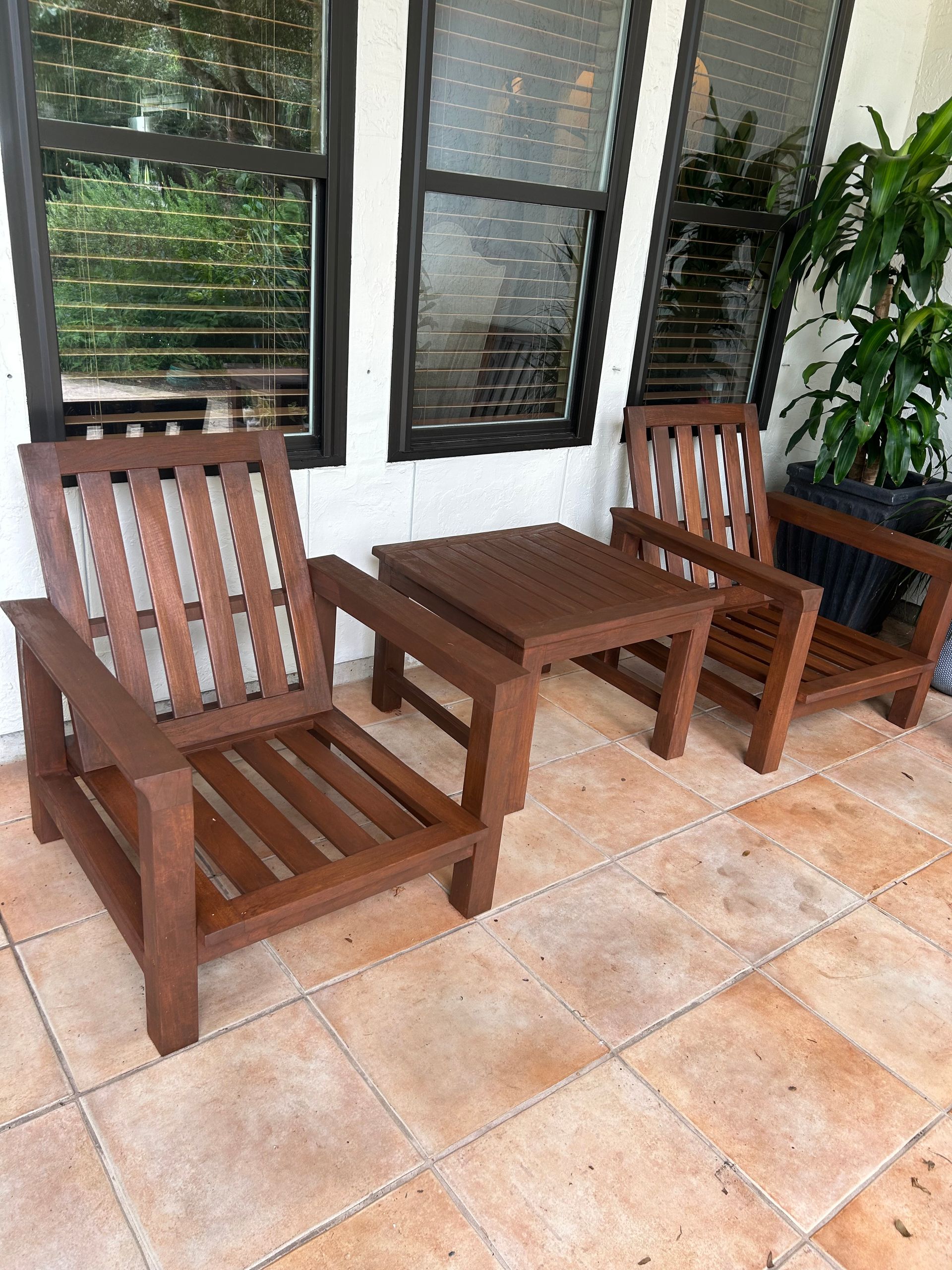 Two brown slatted wooden chairs sit on a tiled patio next to a matching small square table in front of glass doors.