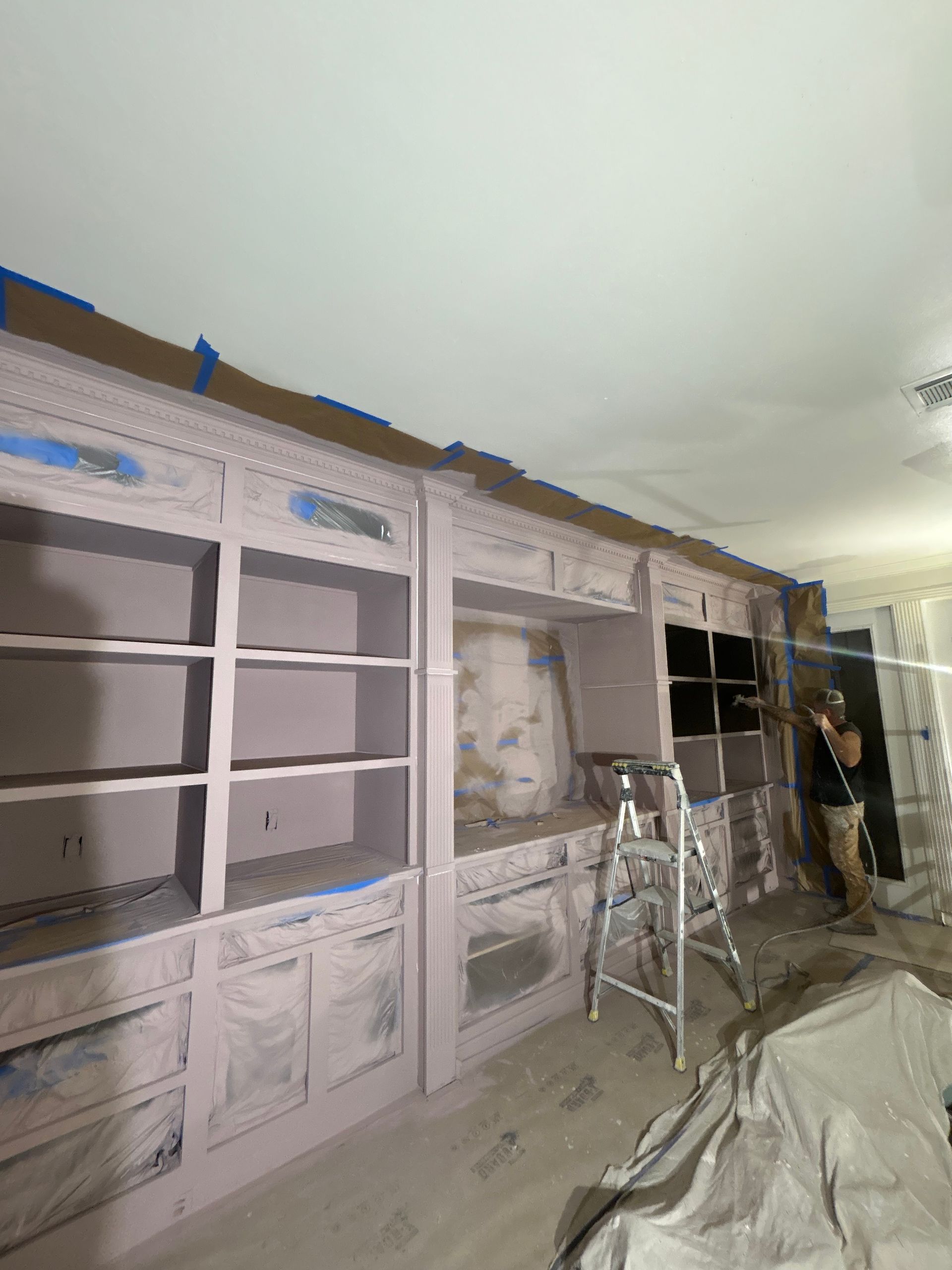 A room under renovation featuring large built-in shelves painted light pink, with a ladder and protective plastic.
