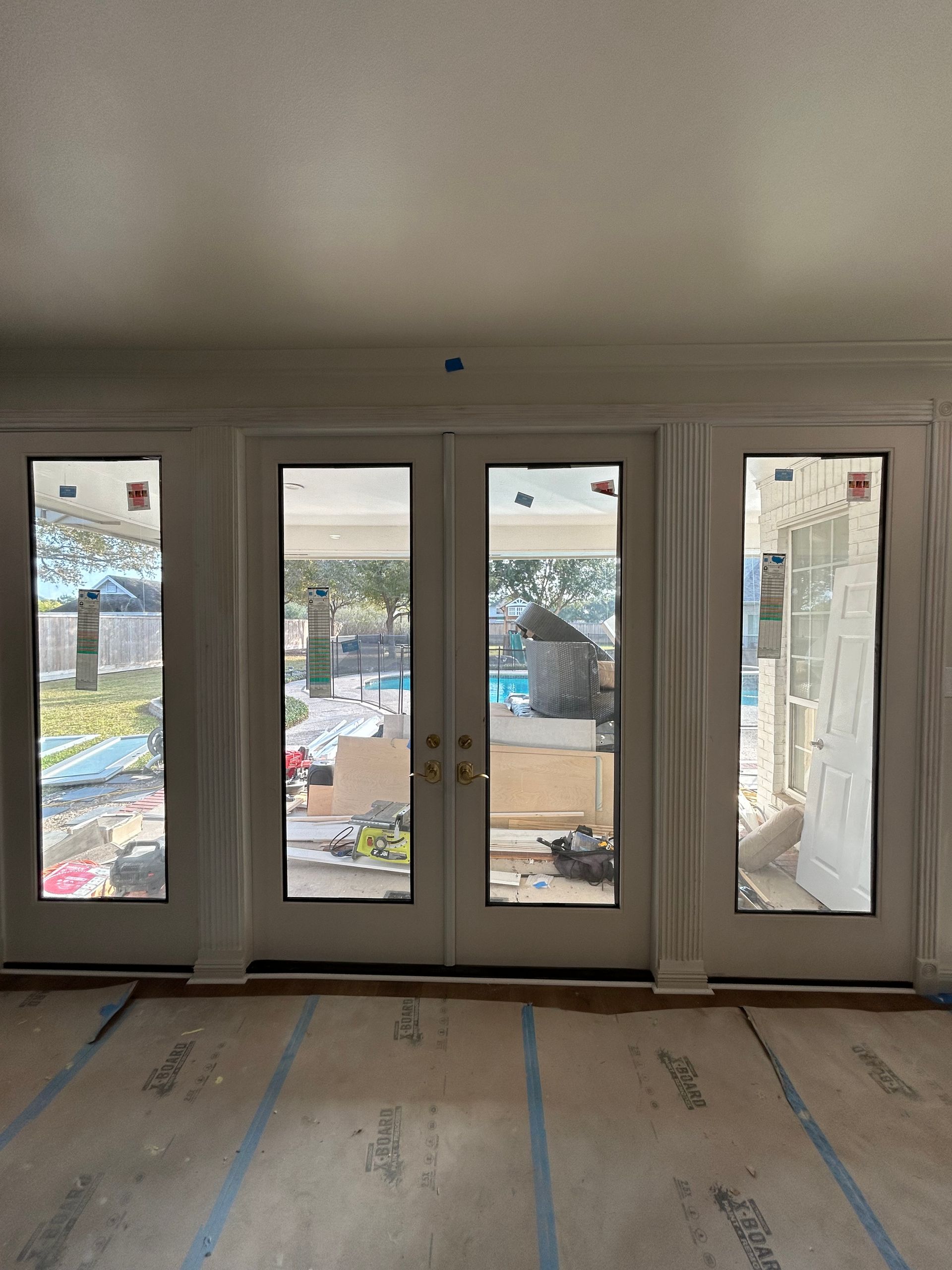 A set of white French doors flanked by sidelights, overlooking an outdoor patio with construction materials on the floor.
