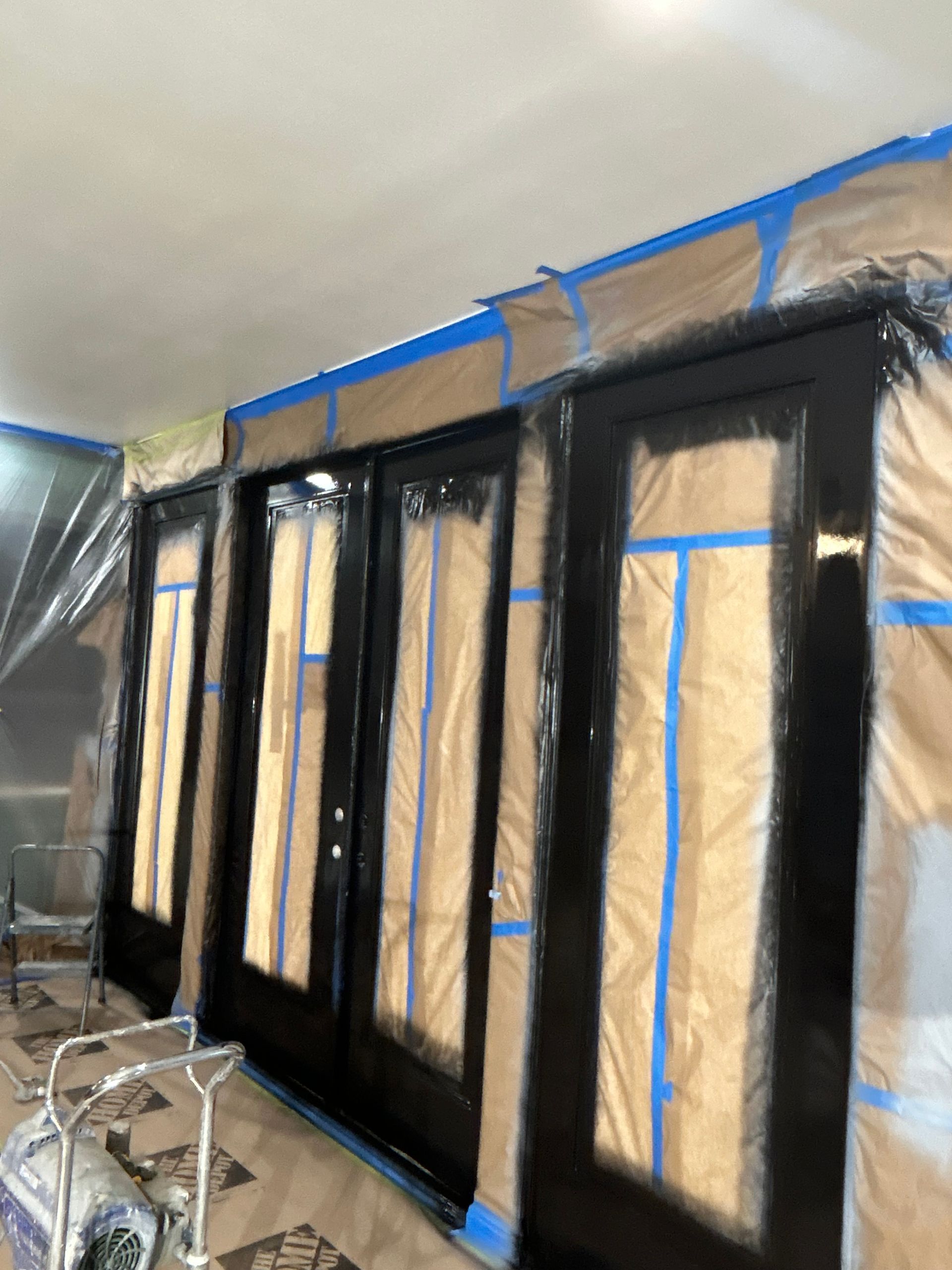 Black french doors prepared for painting, covered with protective plastic sheeting and blue painter's tape.
