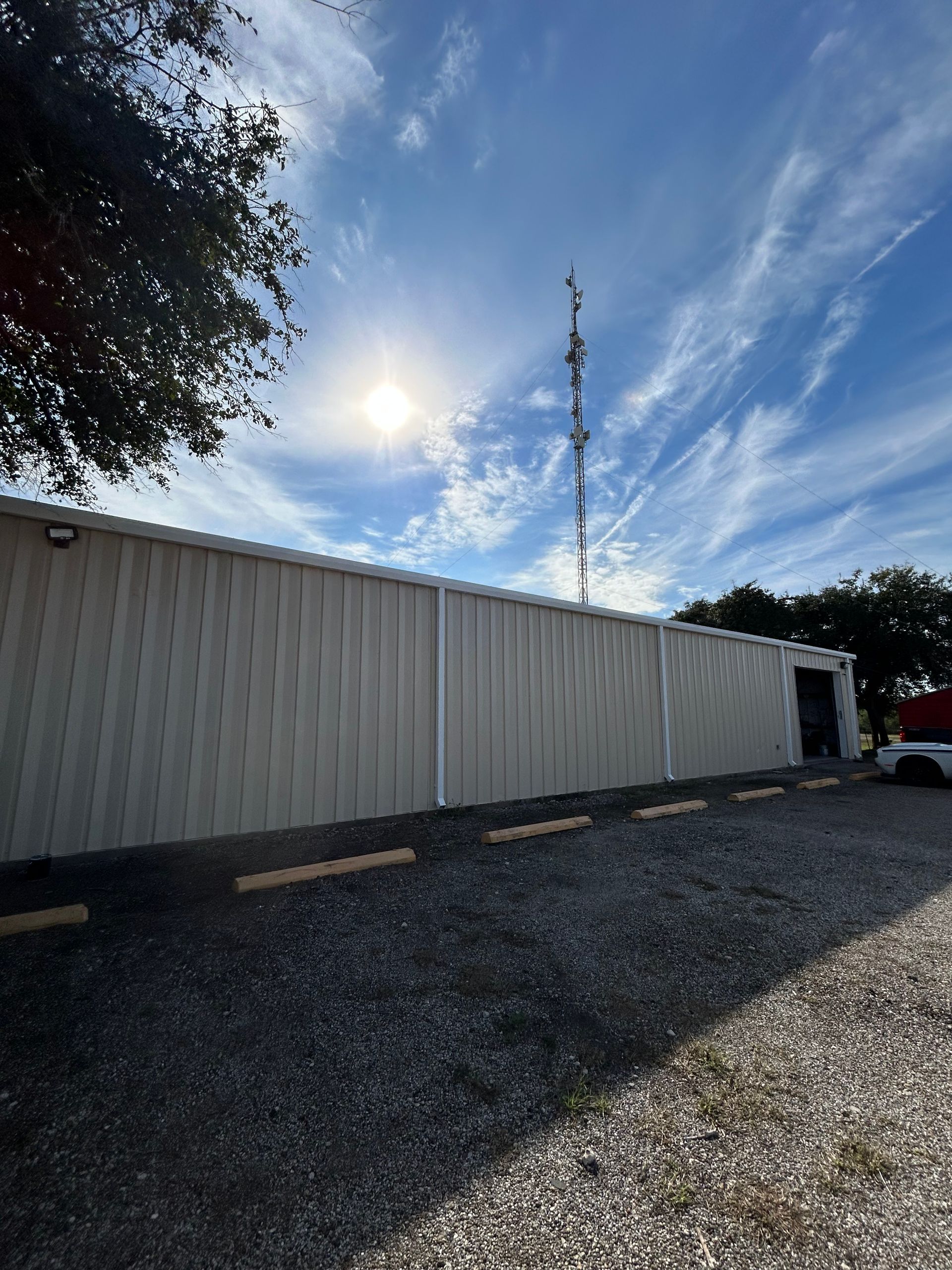 A long, beige metal building sits behind a gravel parking lot under a bright, sunny sky with a tall communications tower.