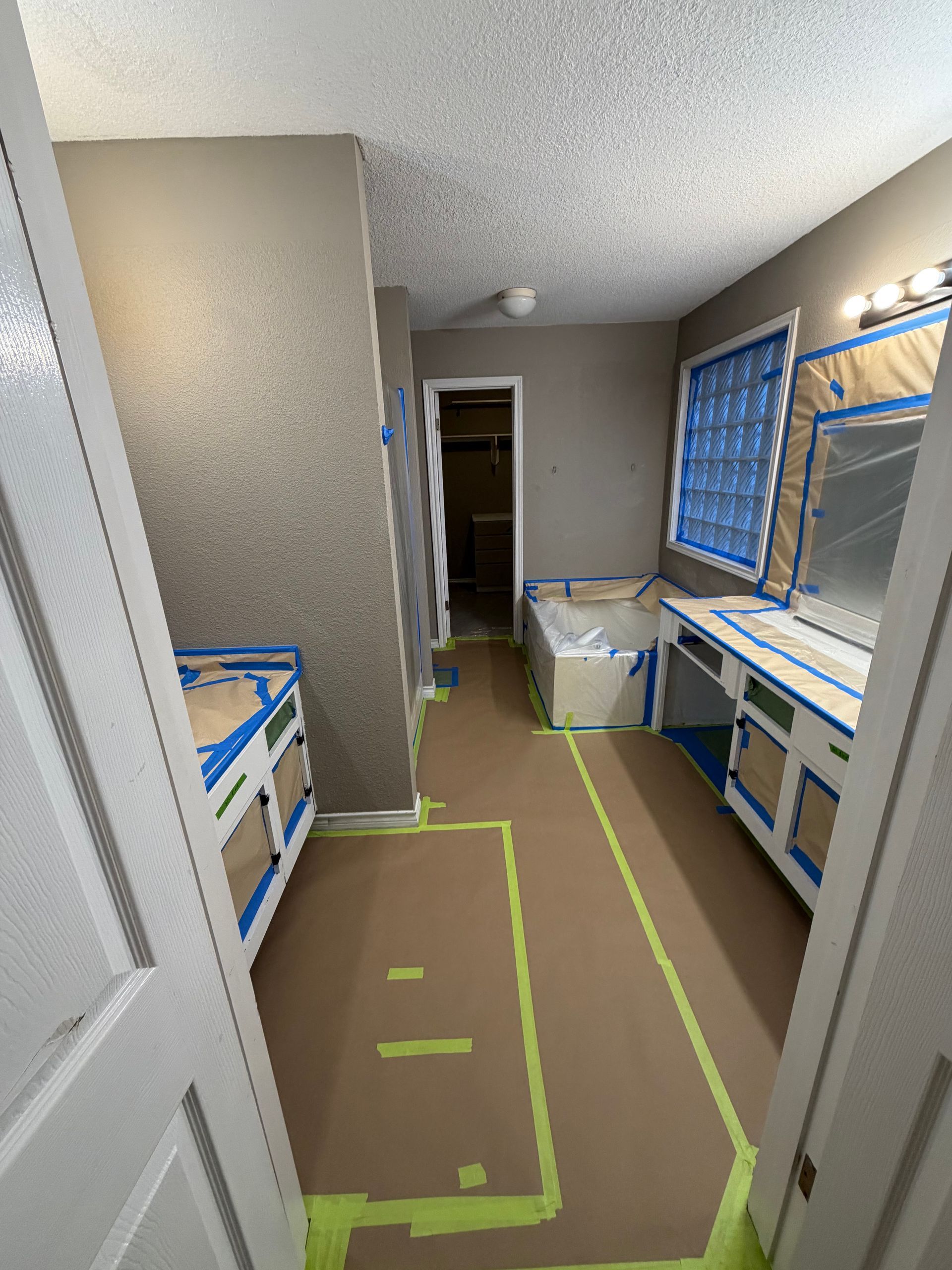 A bathroom undergoing renovation, with cabinets and mirrors taped off, brown paper covering the floor, and tan-painted walls.