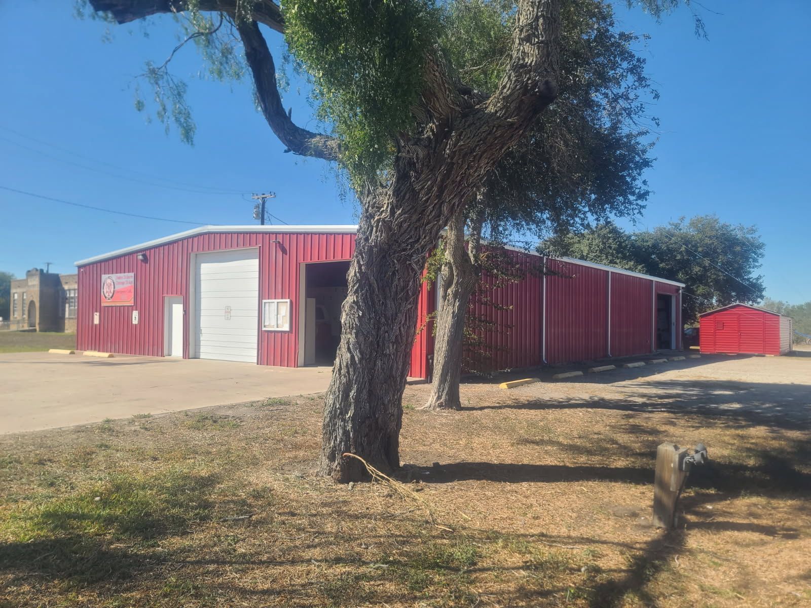 A large red metal warehouse with a white roll-up door stands in a grassy lot under a clear blue sky, partially shaded by trees.