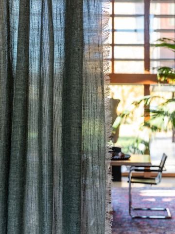 Tenda trasparente accanto a una finestra soleggiata, con una sedia e piante da interno sullo sfondo.