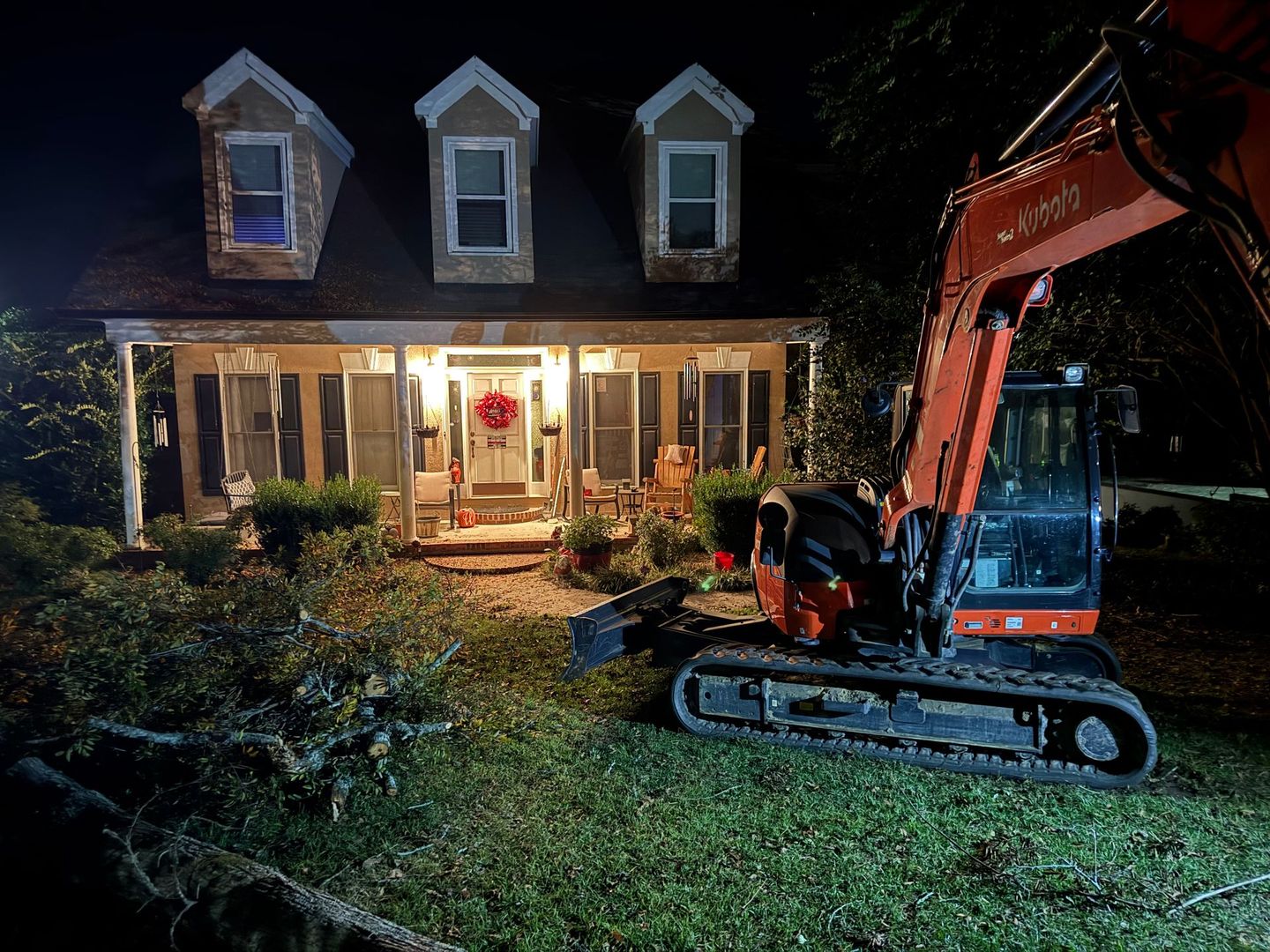 An orange excavator parked in the front yard of a lit house at night.
