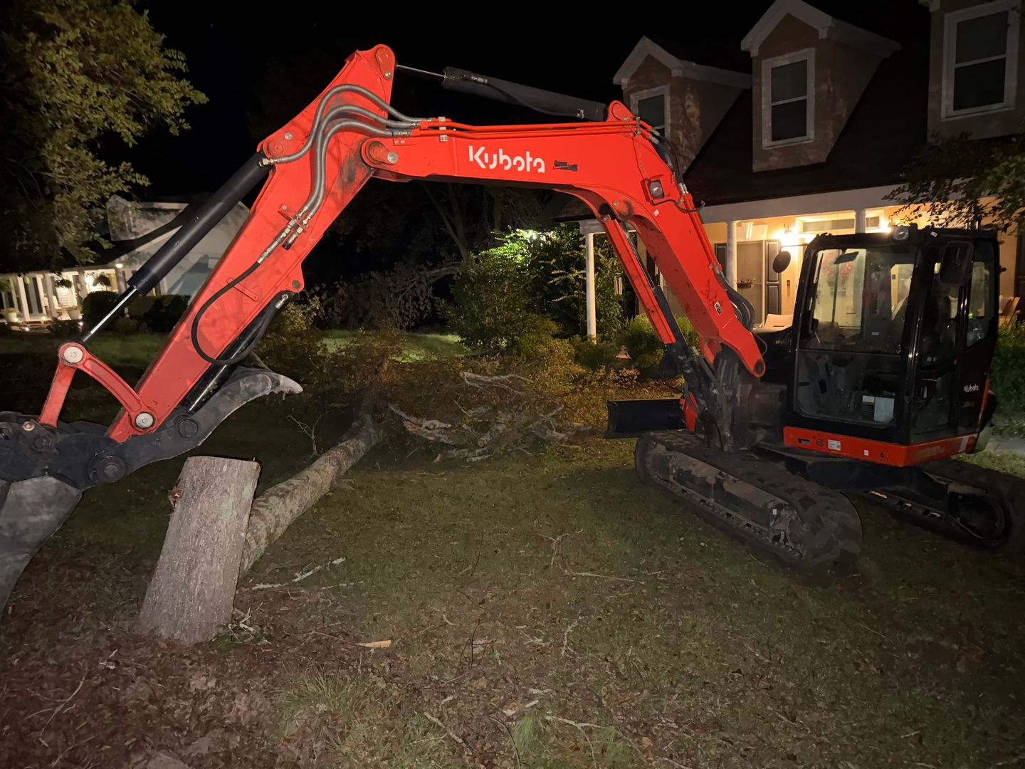 An orange Kubota excavator parked on a lawn at night, positioned near a house and tree debris.