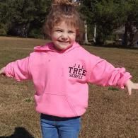 A smiling child in a pink J&L Tree Service hoodie and blue jeans stands in a grassy, sunlit field.
