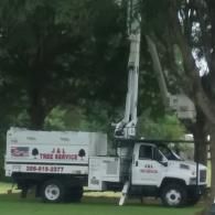 A white J&L Tree Service bucket truck parked on a grass lawn next to trees.