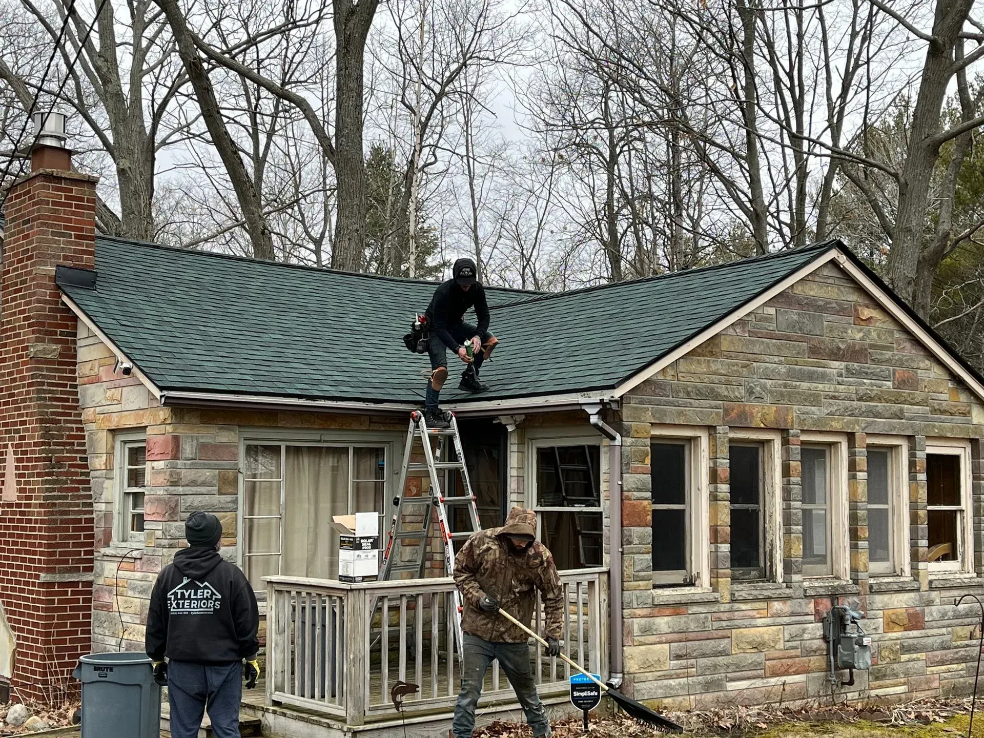 Workers on a roof of a stone house, one on a ladder. Overcast day.
