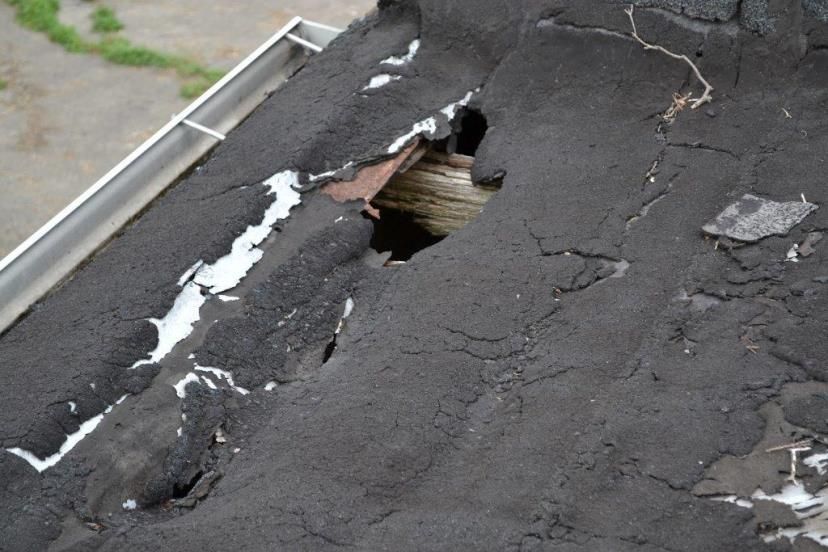 Damaged asphalt roof with large holes, exposing underlying wood and white debris.