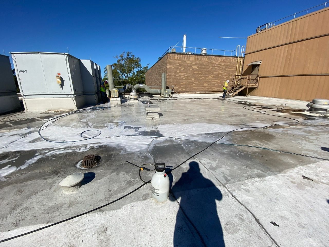 Rooftop with cleaning in progress: person spraying foam on a flat surface, with buildings and equipment visible.