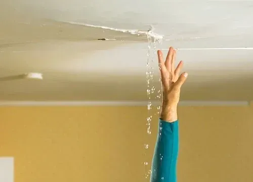 Water leaks from a damaged ceiling onto a raised arm in a room with yellow walls. Arm with blue long sleeve shirt reaching up towards the ceiling.