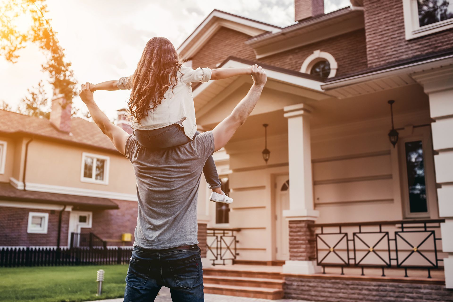 Man carrying a girl on shoulders in front of a house; sunny day.