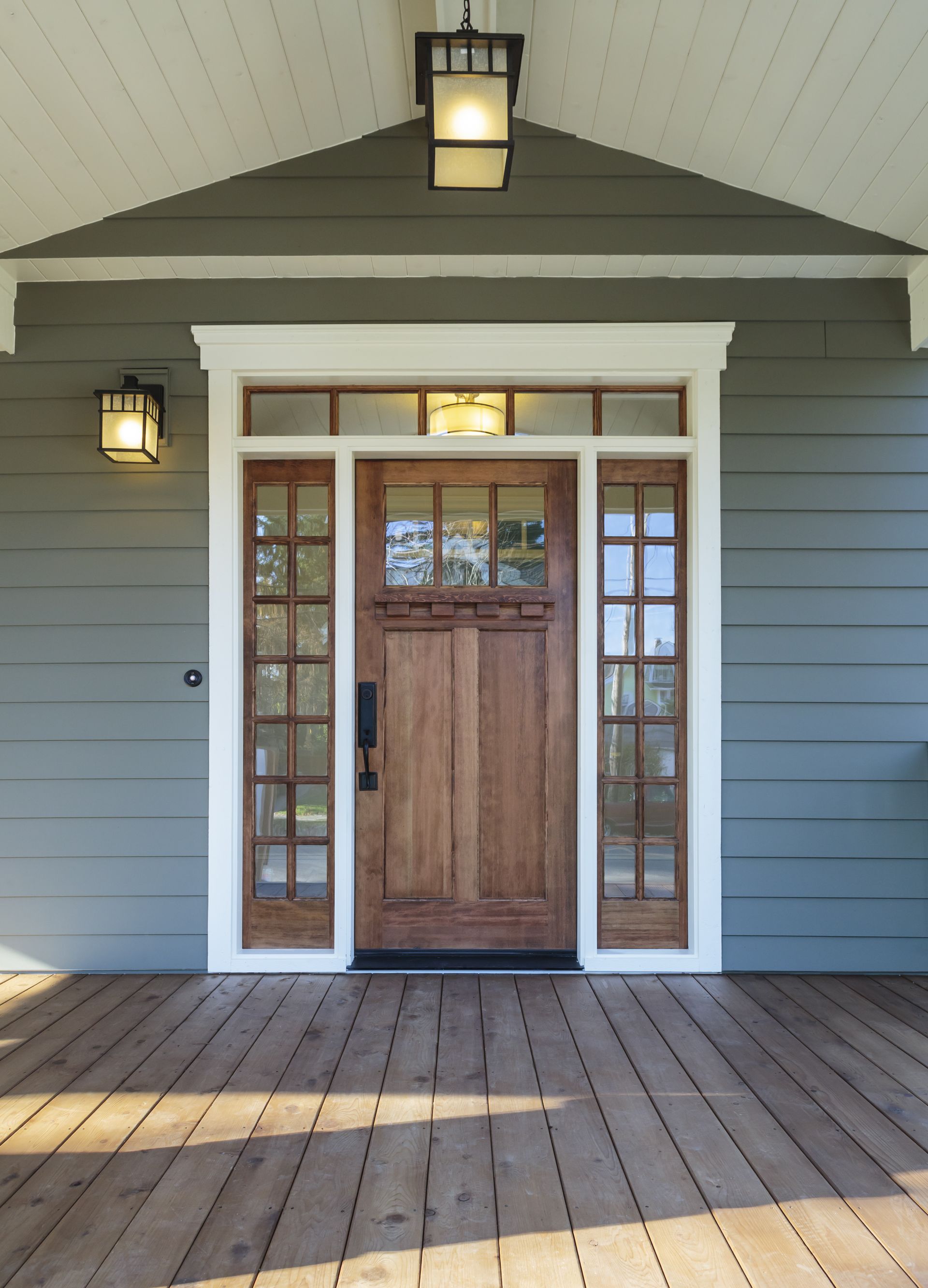 Wooden front door with sidelights and transom, under a porch roof, flanked by sconce lights, wood porch floor, gray siding.