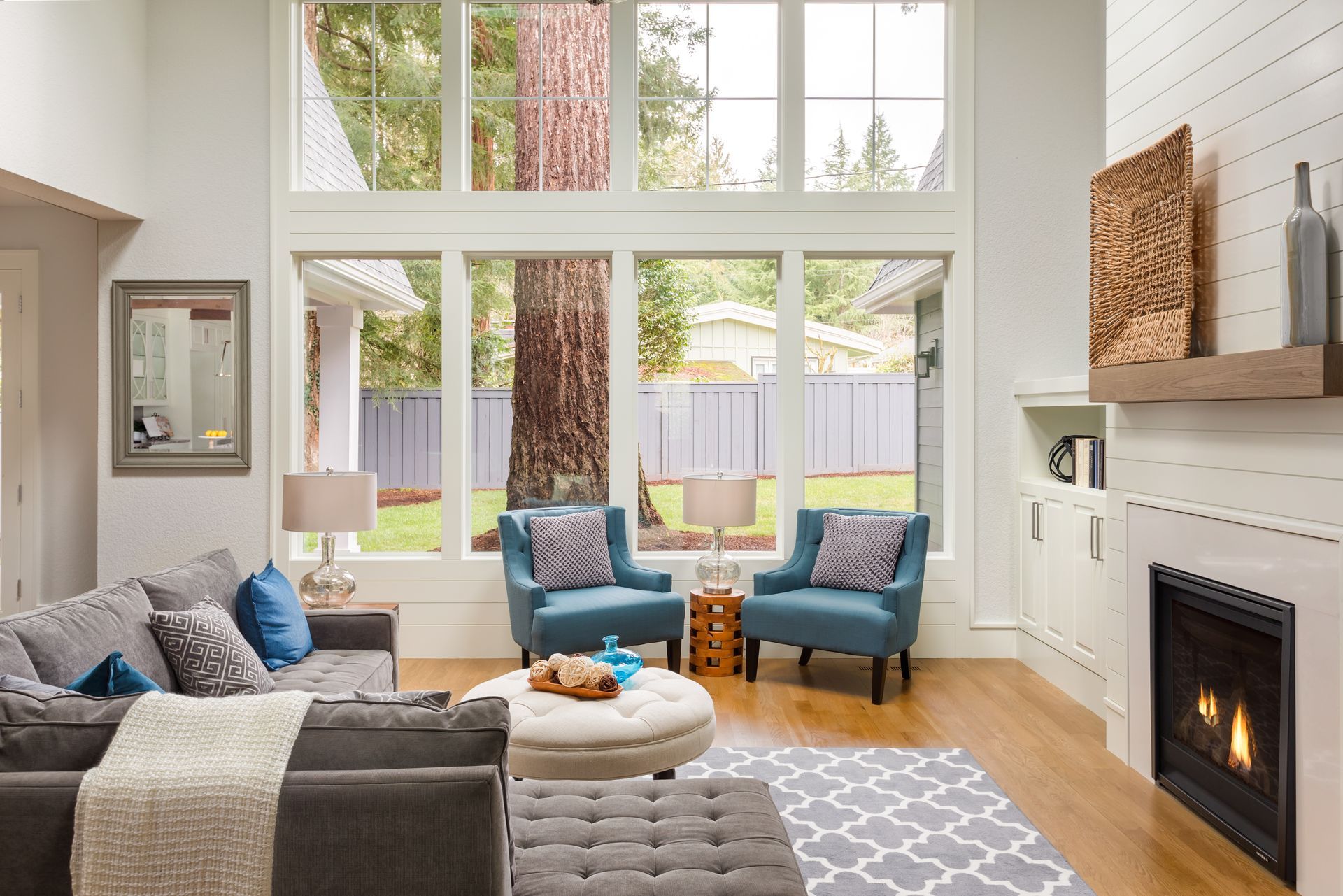 Living room with large windows, blue chairs, gray sofa, fireplace, and hardwood floors.