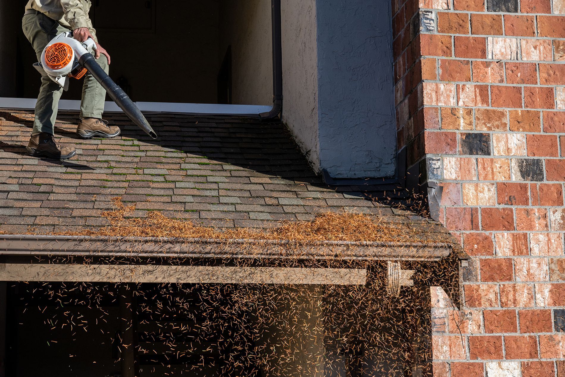 Person using a leaf blower on a roof, blowing debris toward a gutter near a brick building.