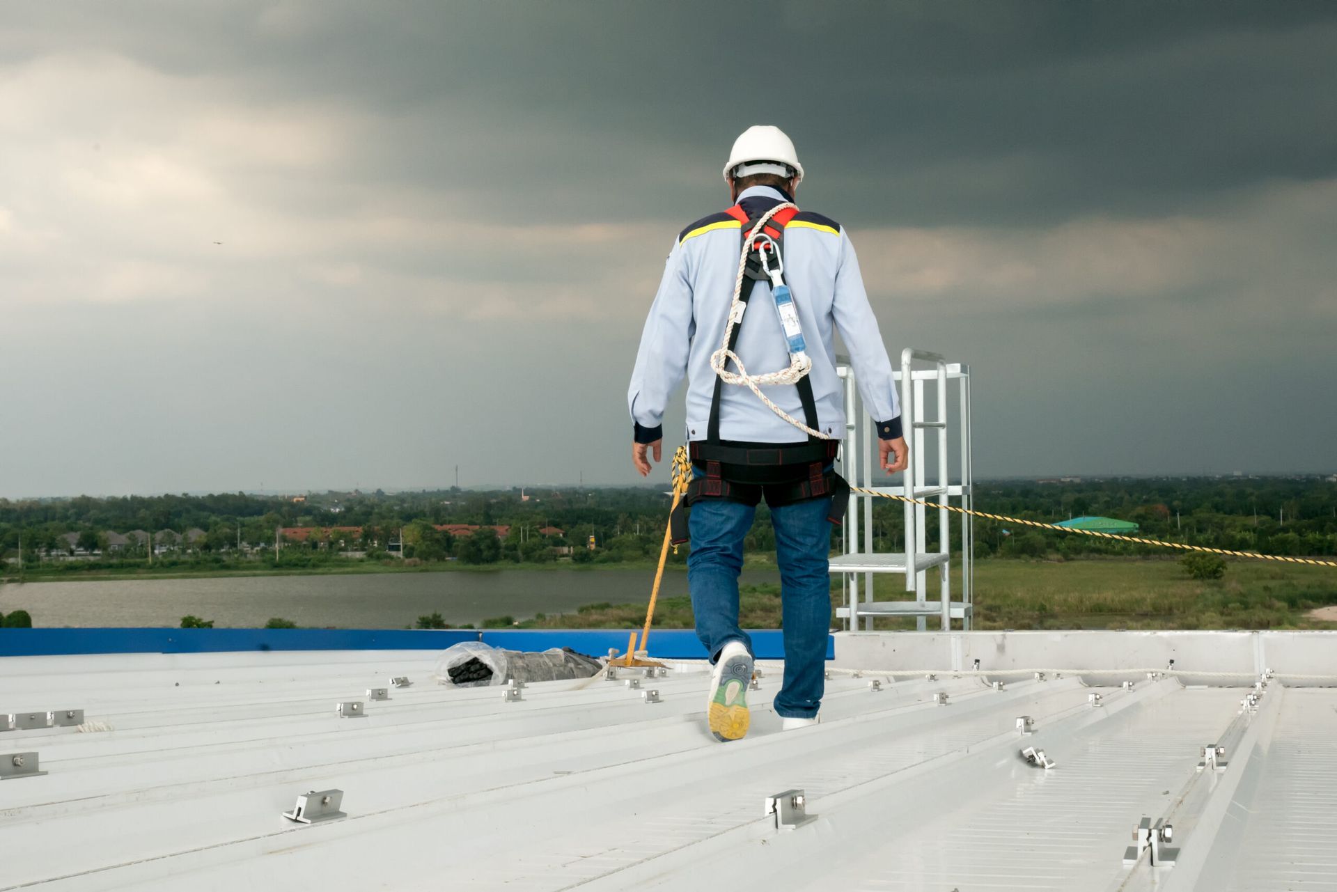 Person in safety harness and white helmet on a rooftop, walking toward a cloudy sky.