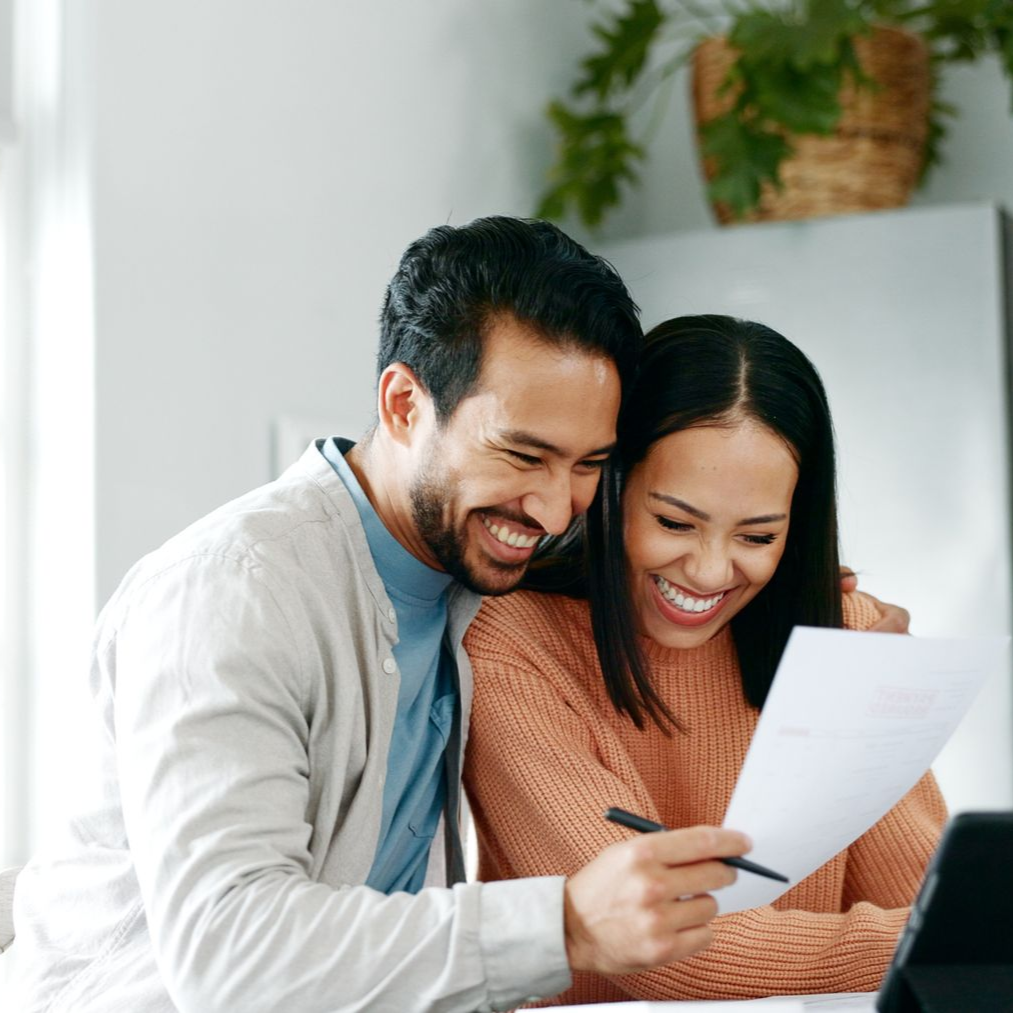 Couple smiling, looking at document. One has arm around the other. Interior setting, natural light.