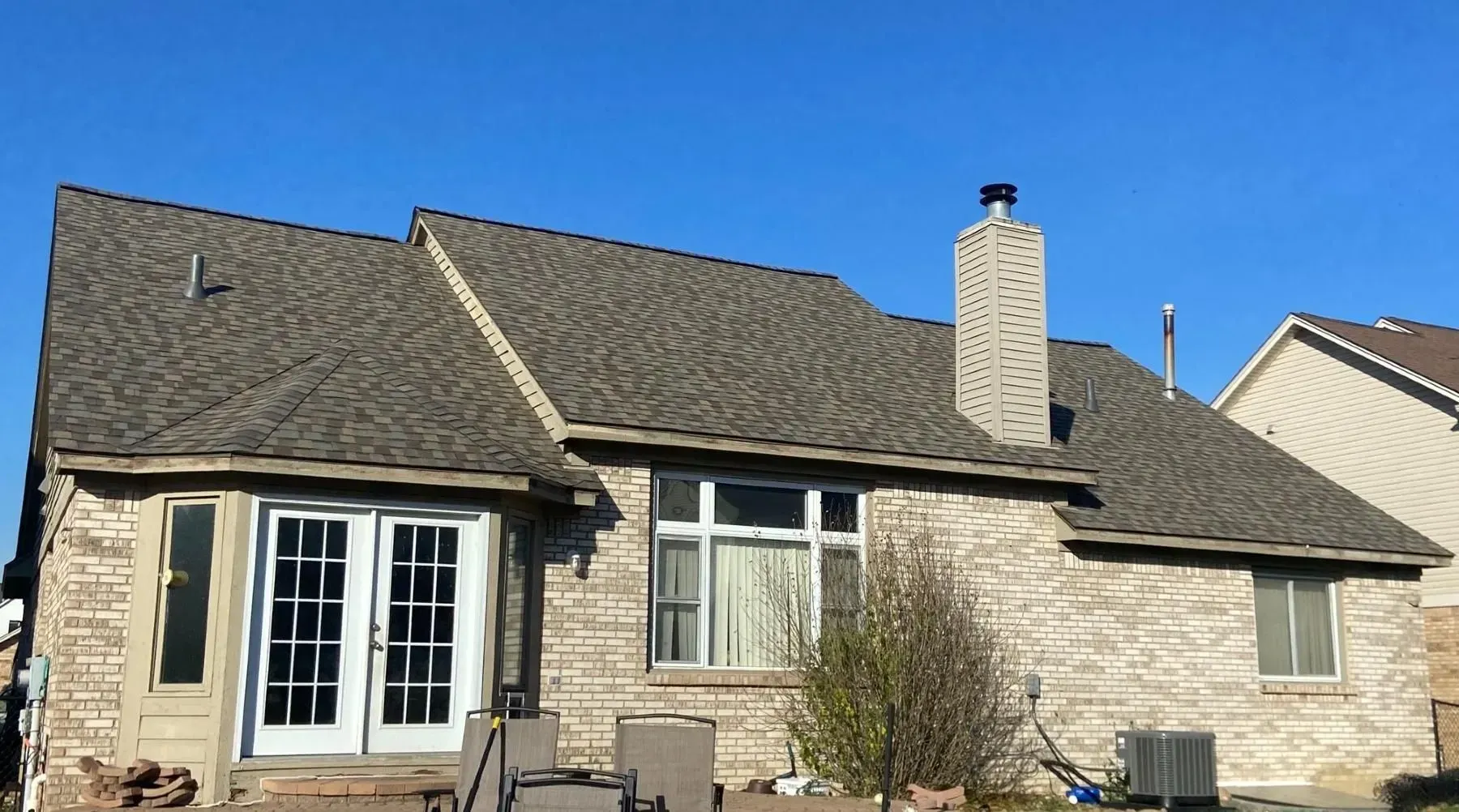 Backyard view of a brick house with a brown shingled roof, windows, and a chimney against a blue sky.
