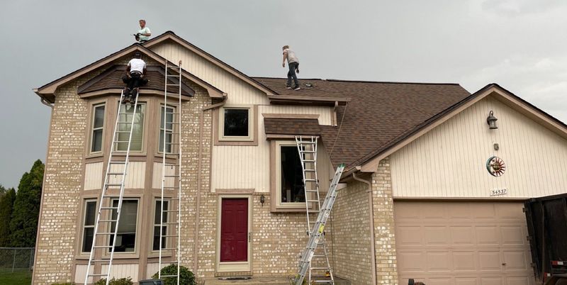 Two people on a roof working. Ladders, light brick house, dark roof, cloudy sky.