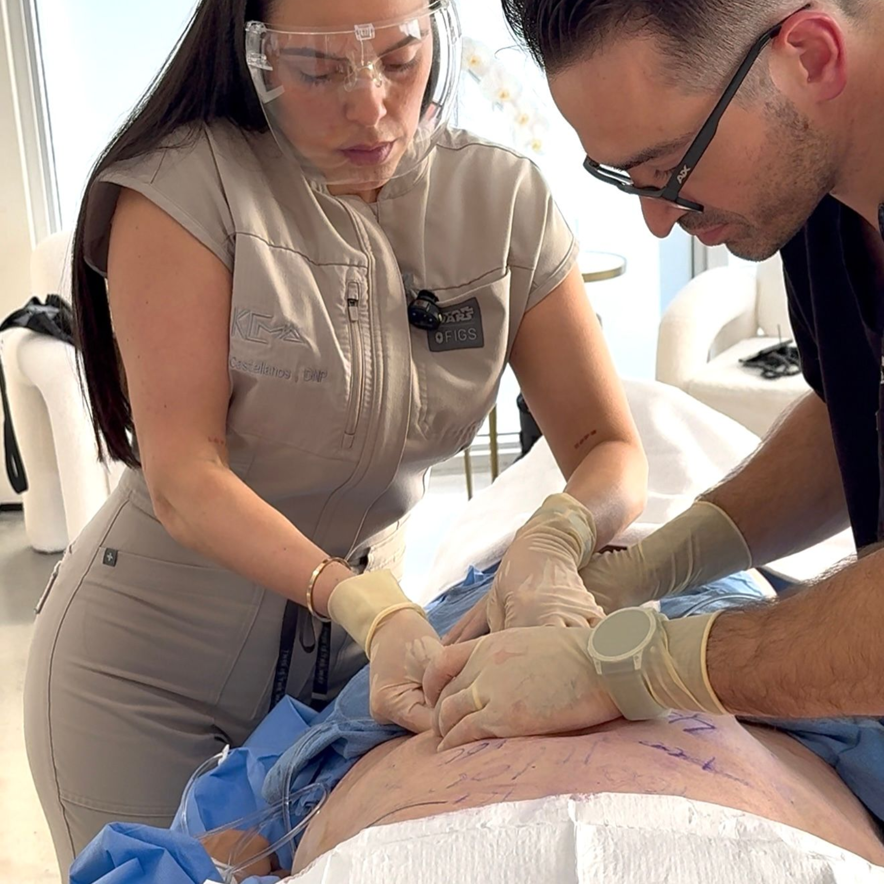 Two medical professionals in scrubs and gloves performing a procedure on a patient's abdomen in a clinical setting.