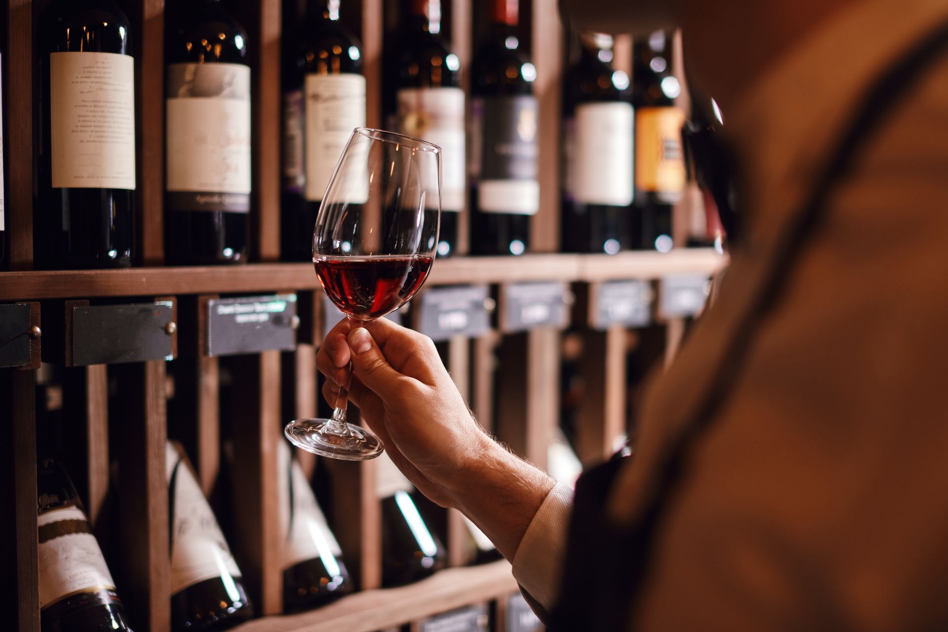 Bartender or male cavist standing near the shelves of wine bottles holds a glass of wine, looks at tint and smells flavor of wine in glass.