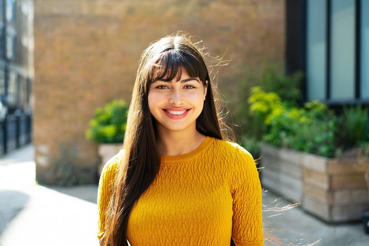 Woman with long dark hair and bangs smiles, wearing a yellow sweater, outdoors in front of a brick wall.