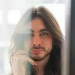 Man with long brown hair and a small beard, gazing at the viewer, with a thoughtful expression.