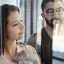 Woman and man looking at something, possibly through glass. Man wears glasses and a beard.