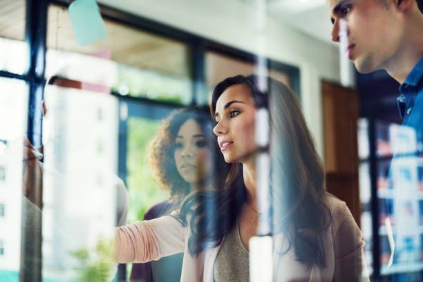 Three people looking at sticky notes on a glass wall; brainstorming in an office.