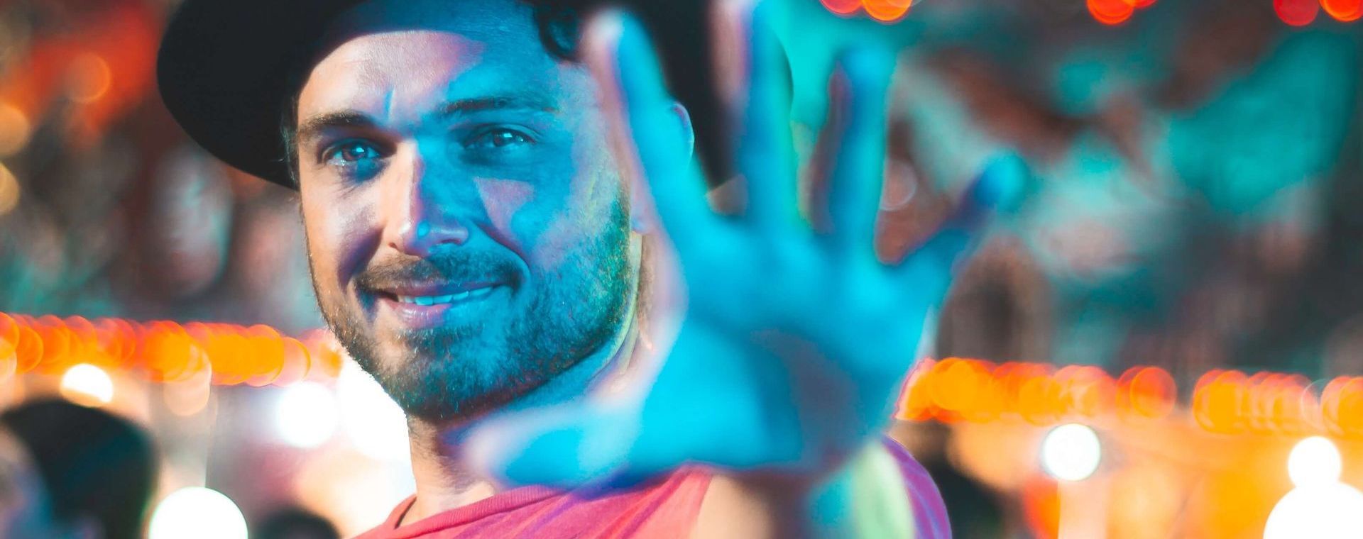 Man in a hat with hand up, smiling at a colourful event with bokeh lights.