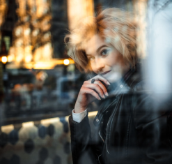Woman with blonde hair, leaning against a window, gazing thoughtfully. Wearing black jacket, cafe setting.