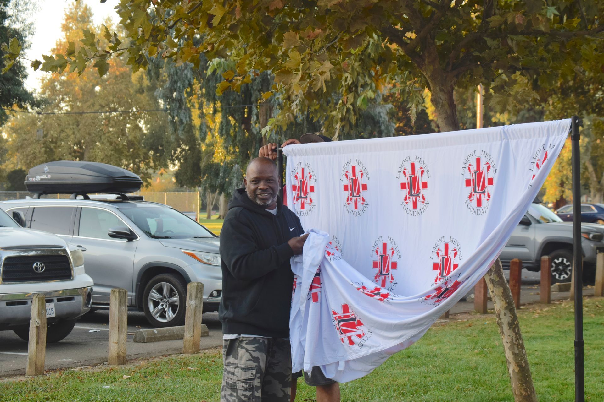 Man holding a white backdrop with red symbols, next to cars and trees outdoors.