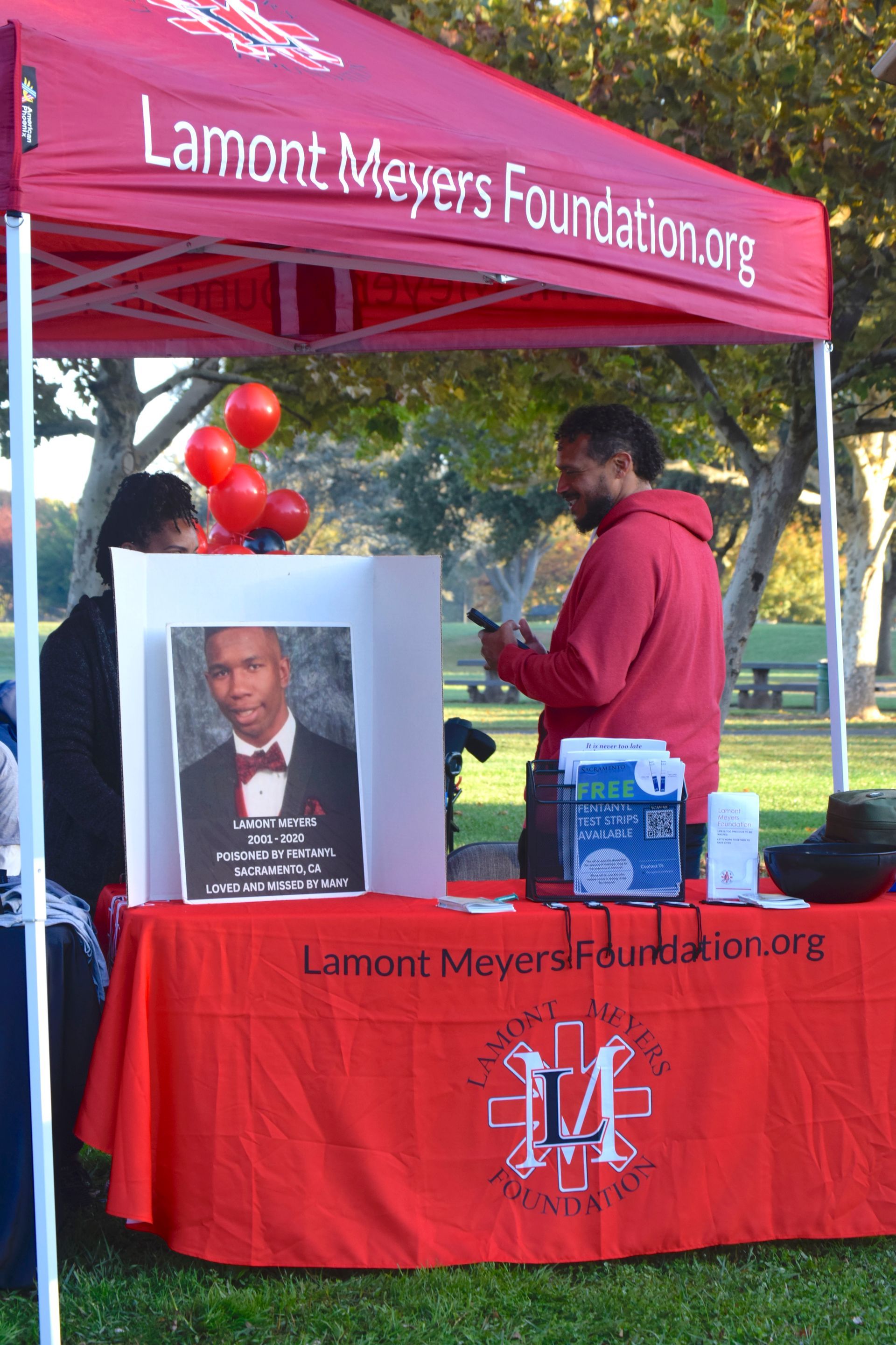 A booth for the Lamont Meyers Foundation at an outdoor event, displaying a portrait and information. A man looks at a phone.