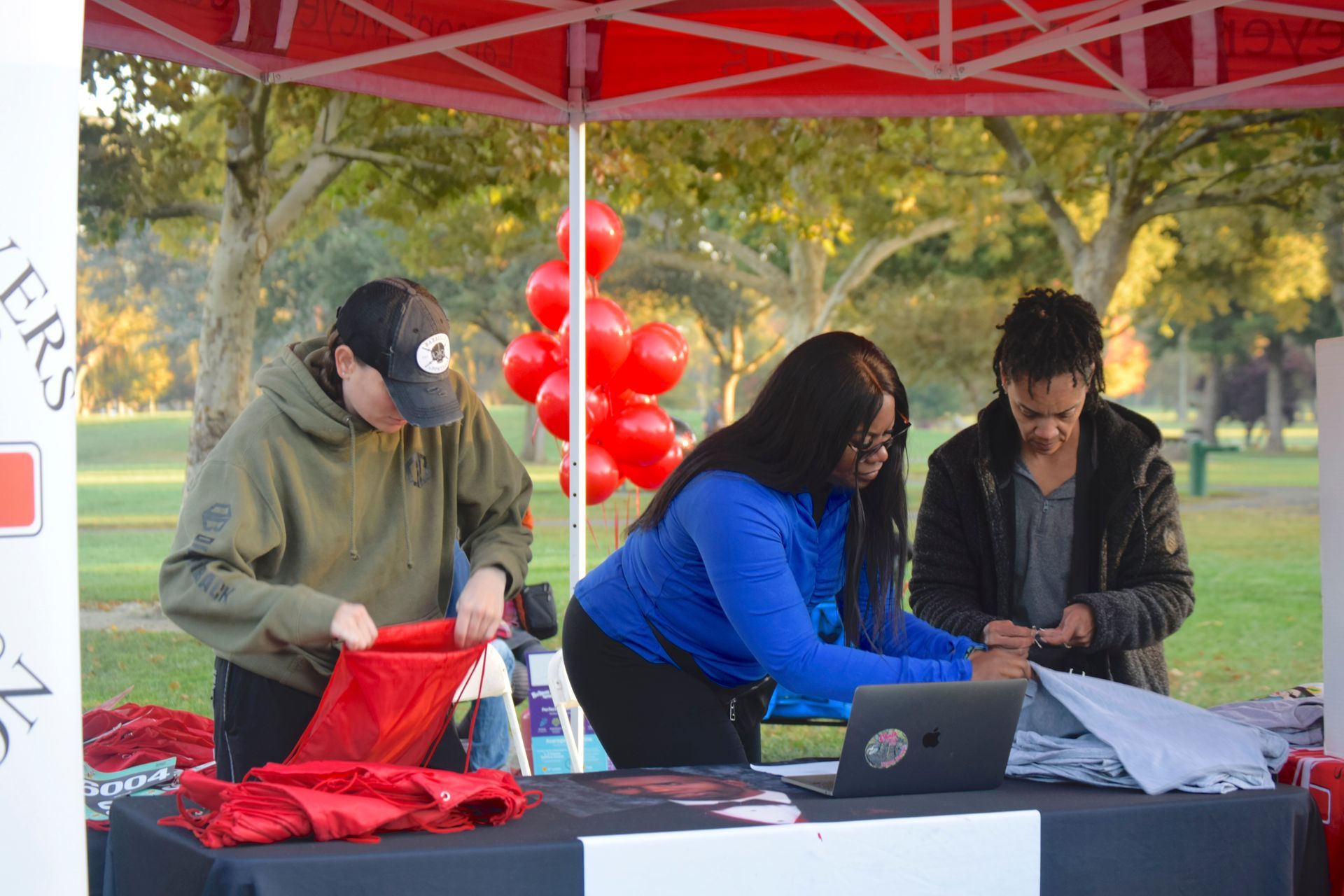 People at a red tent table, setting up items. Balloons are in the background, a park setting.