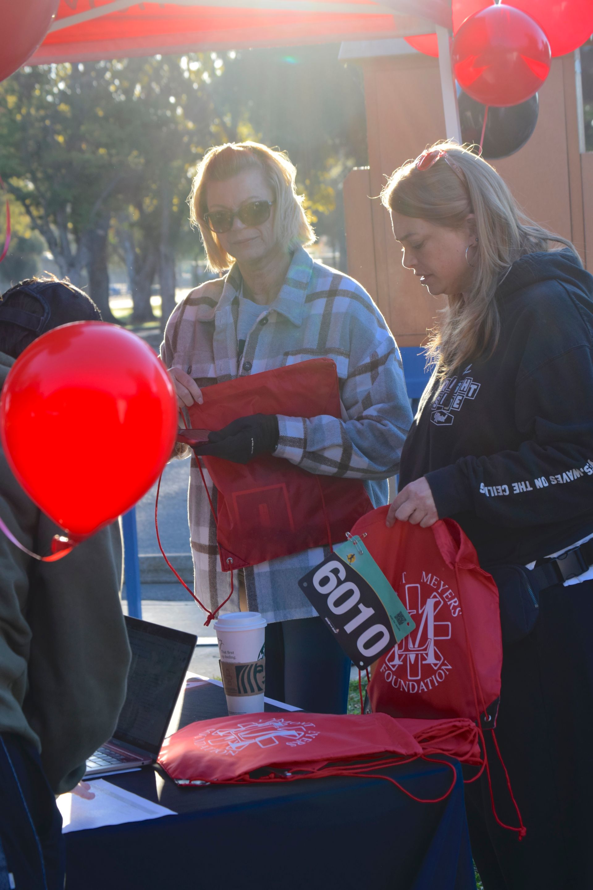 People at an outdoor event, examining red bags at a table. Red balloons in the background.
