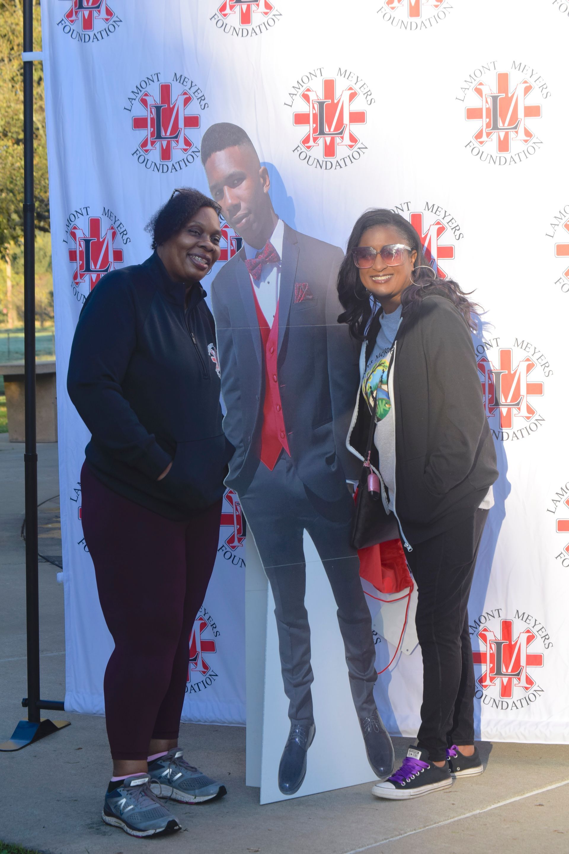 Two women pose beside a cardboard cutout of a man against a banner. Outdoor setting, sunny day.