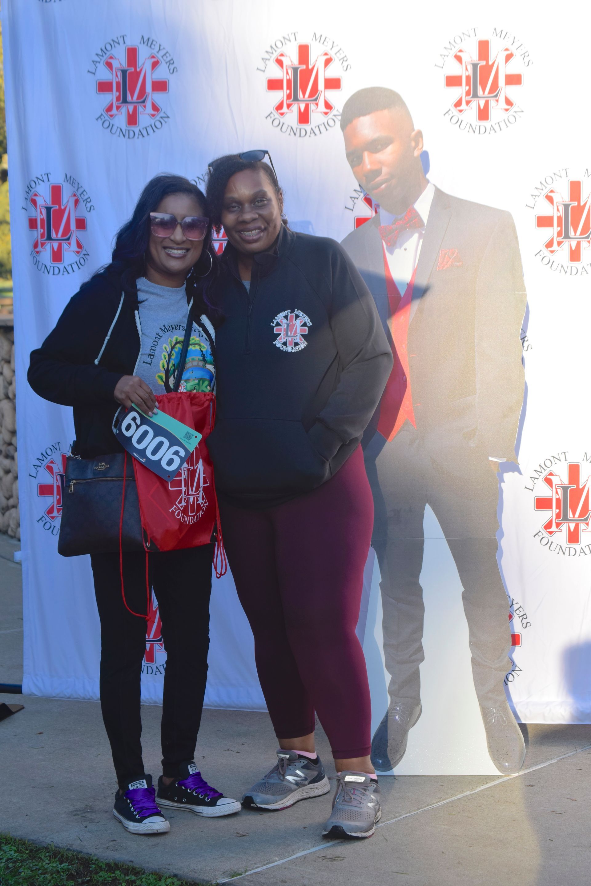 Two women pose beside a cardboard cutout of a man at an outdoor event. One woman holds a bag and race number.