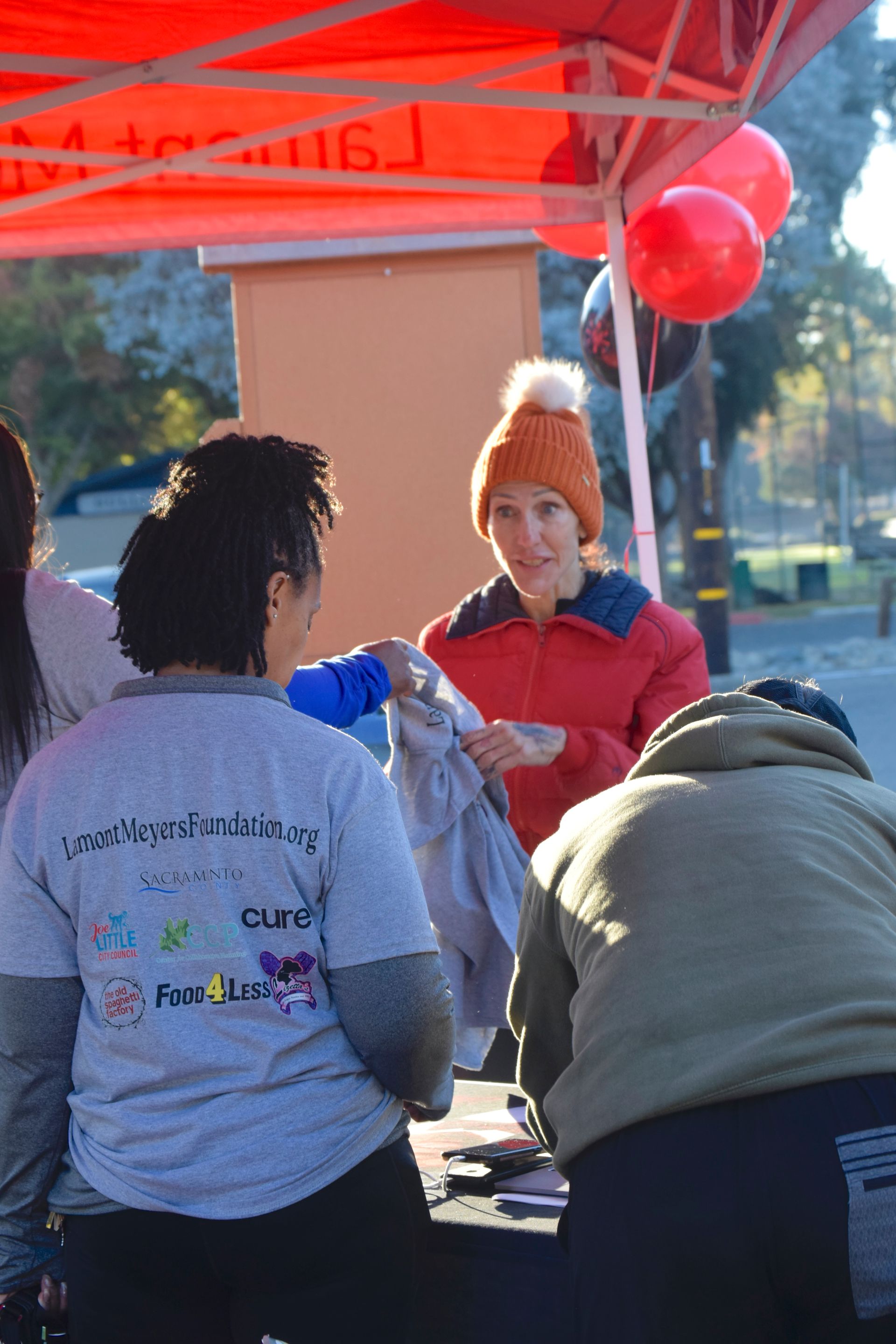 People at an outdoor event, one handing a bag. A woman in an orange hat, red jacket, and balloons in the background.