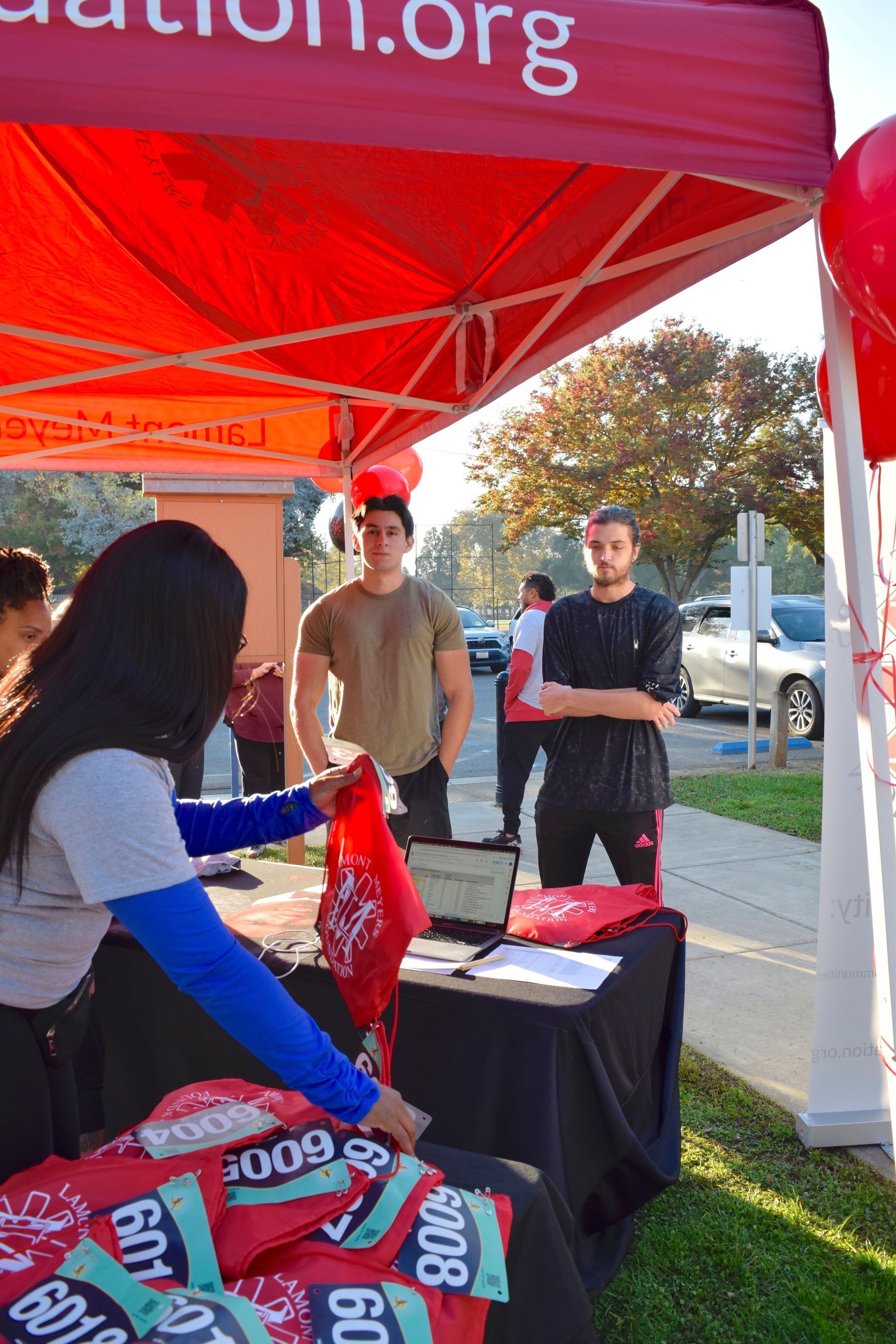 People at a registration table under a red canopy. A woman hands a red item to two men.