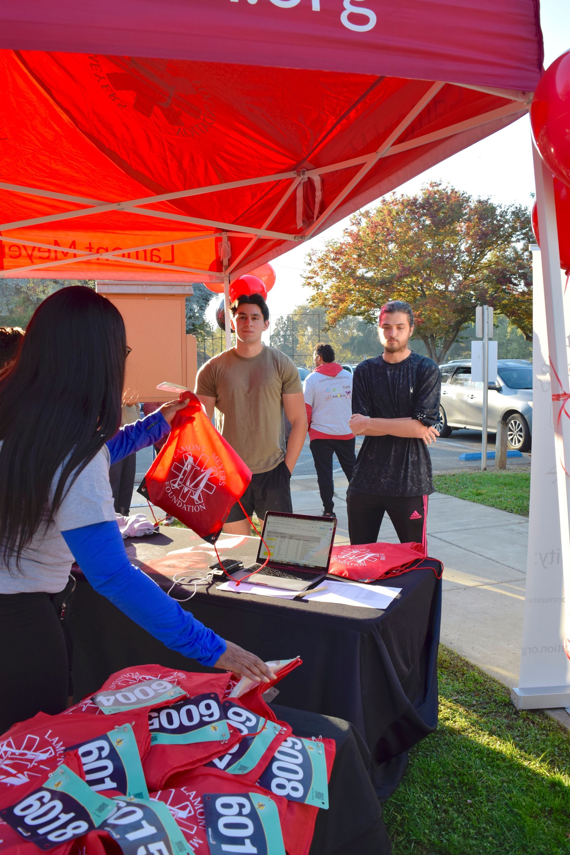People at a table under a red canopy, handing out bags and race bibs.